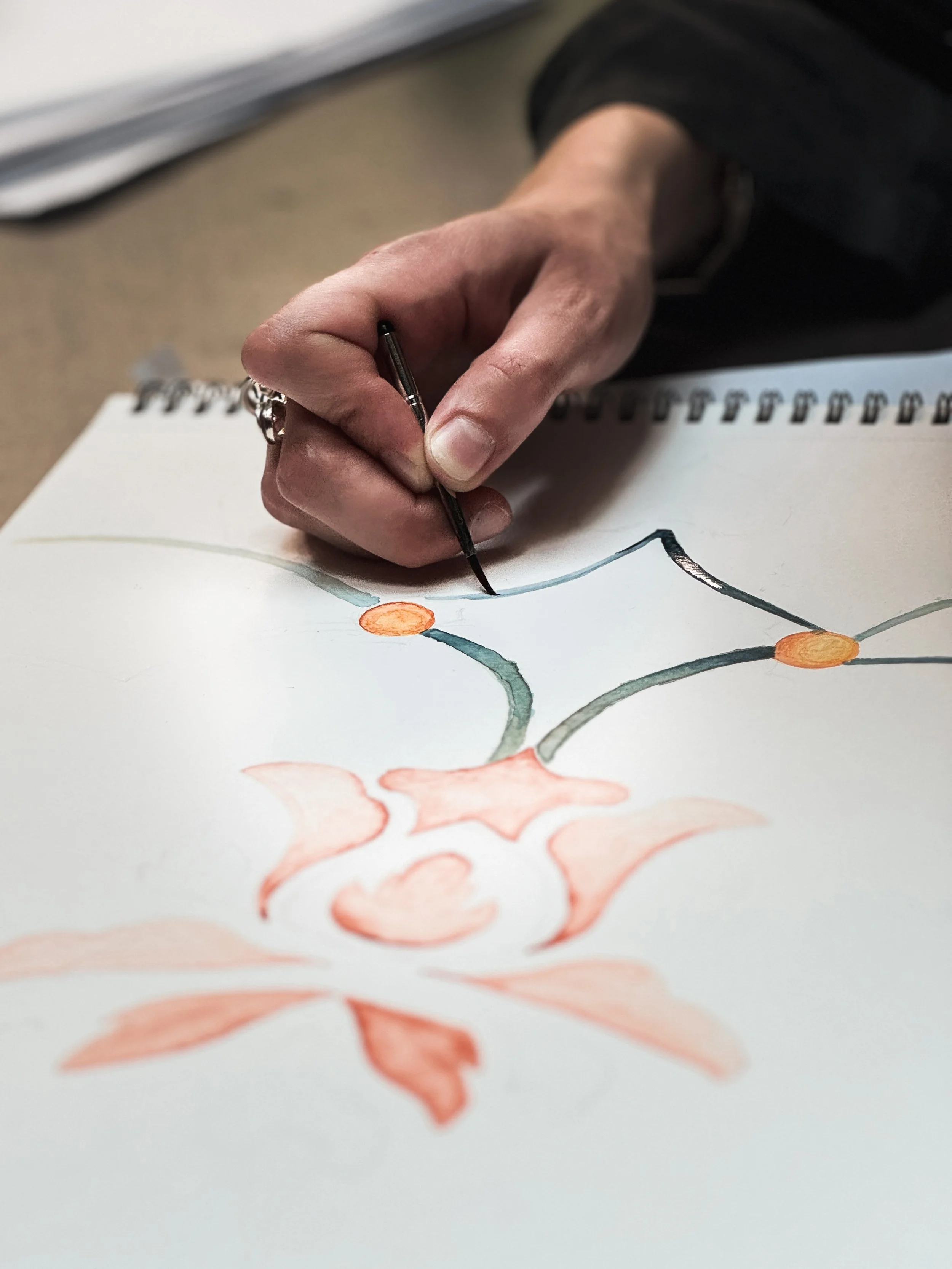A person drawing with a fine pen on a sheet of paper with a watercolor illustration of abstract, tree-like shapes in orange and gray.