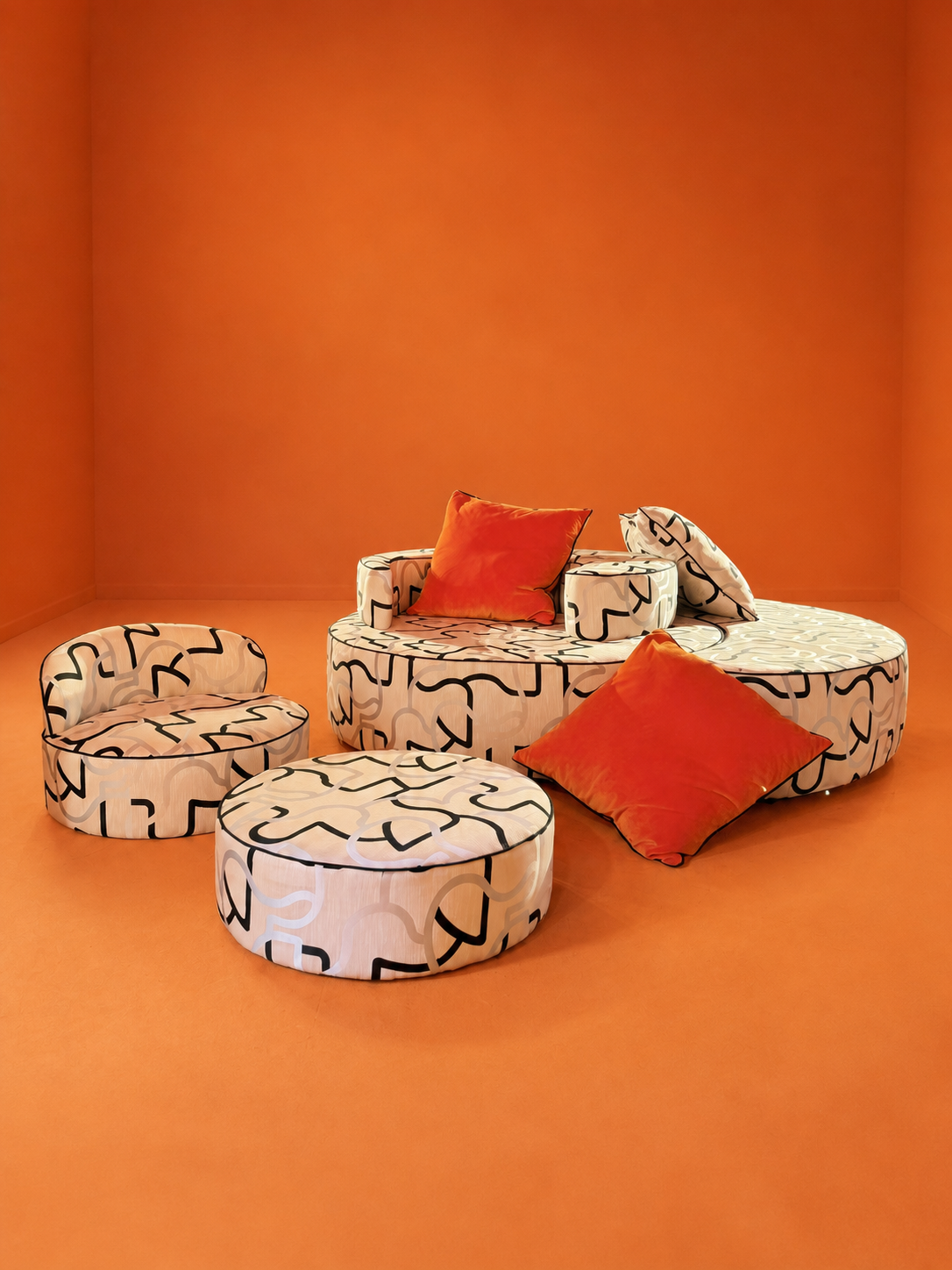 Room with orange walls and floor, featuring abstract-patterned seating including a round sofa, ottomans, and cushions, primarily in cream and black with orange cushions.