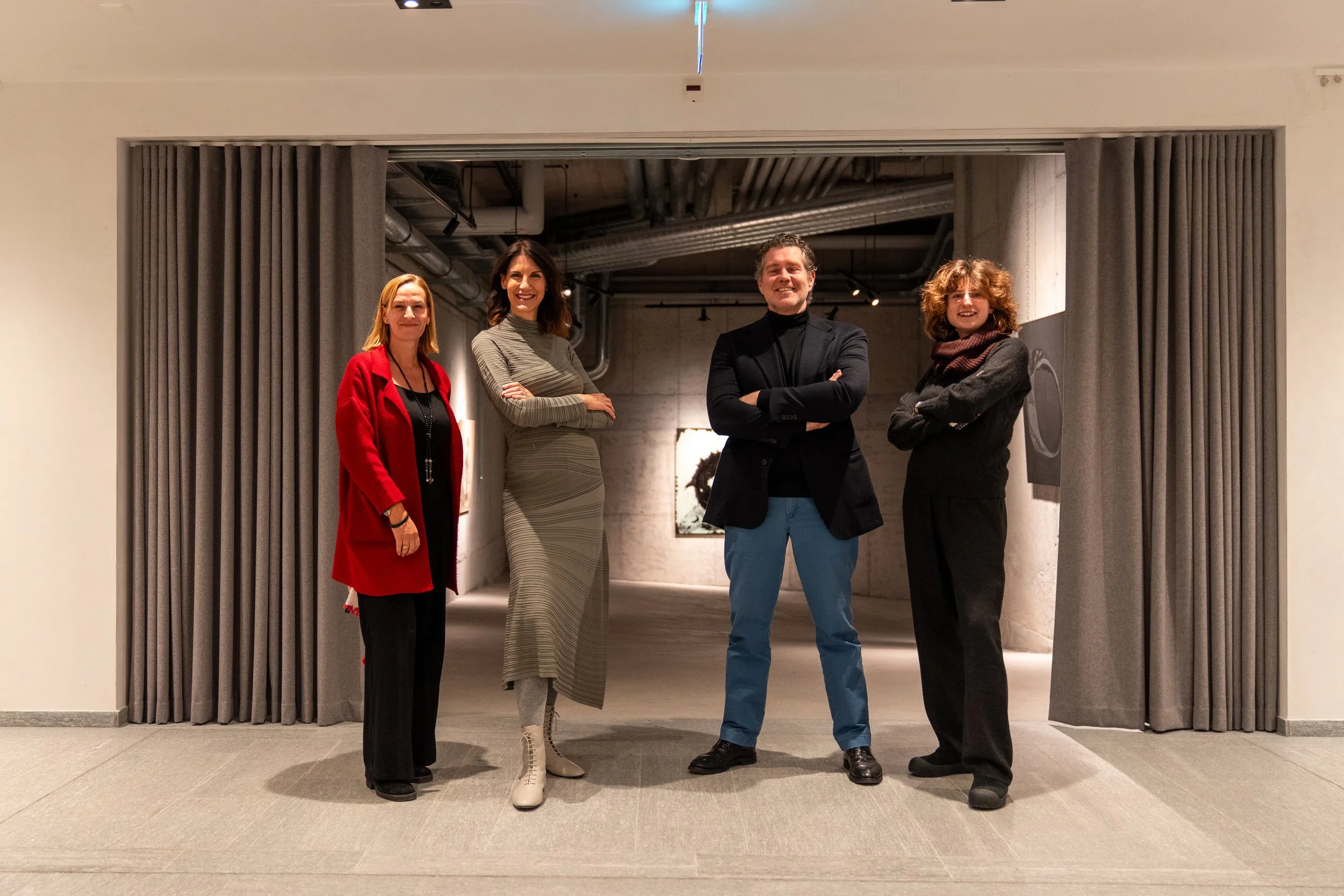 Four people standing in a gallery, posing for a photo. They are standing in front of an open doorway with grey curtains, and some artwork is visible in the background.