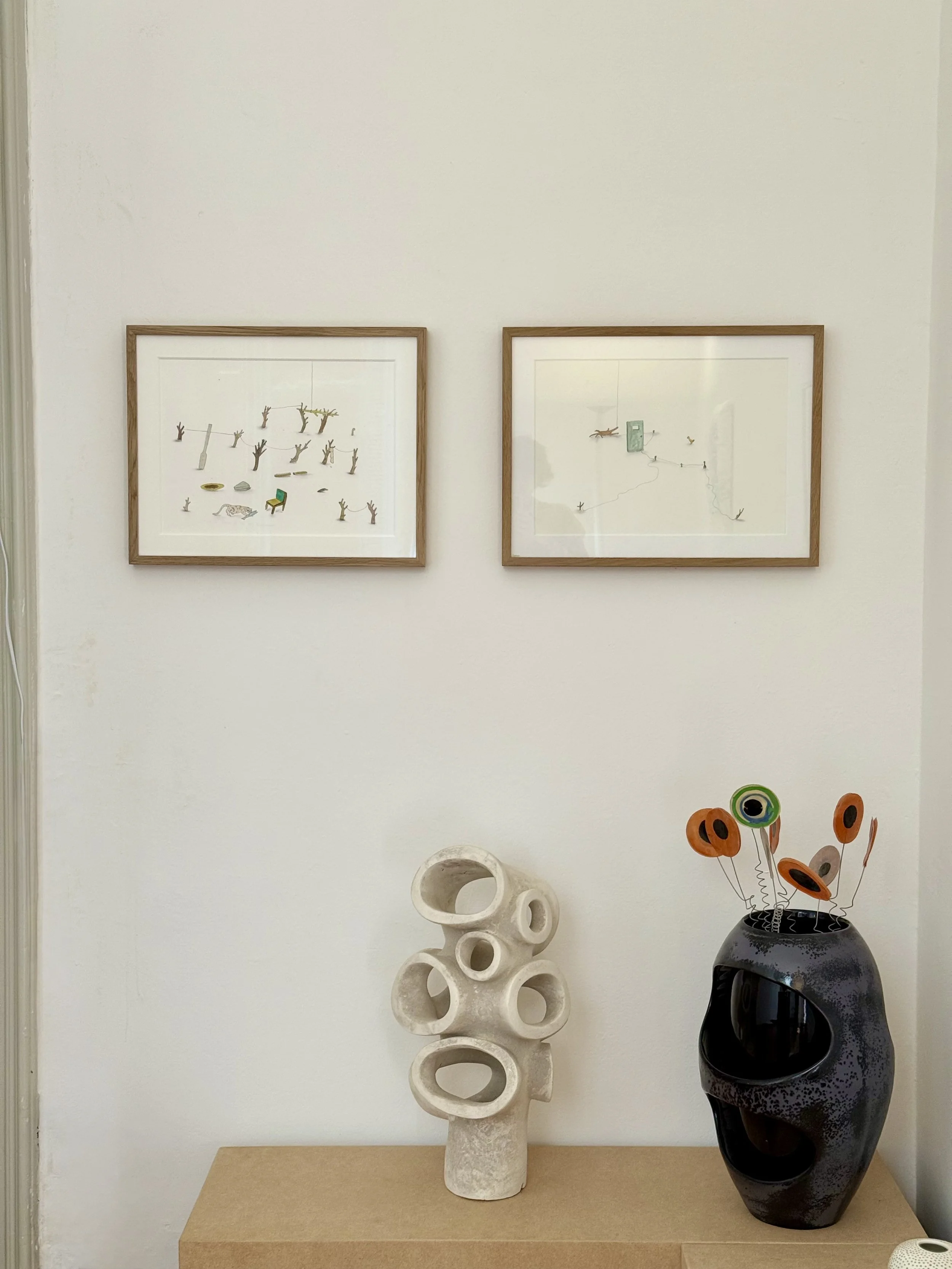 Decorative interior display with two framed art pieces on white wall, white sculptural object with circular holes, black vase with abstract orange and black flower sculpture.