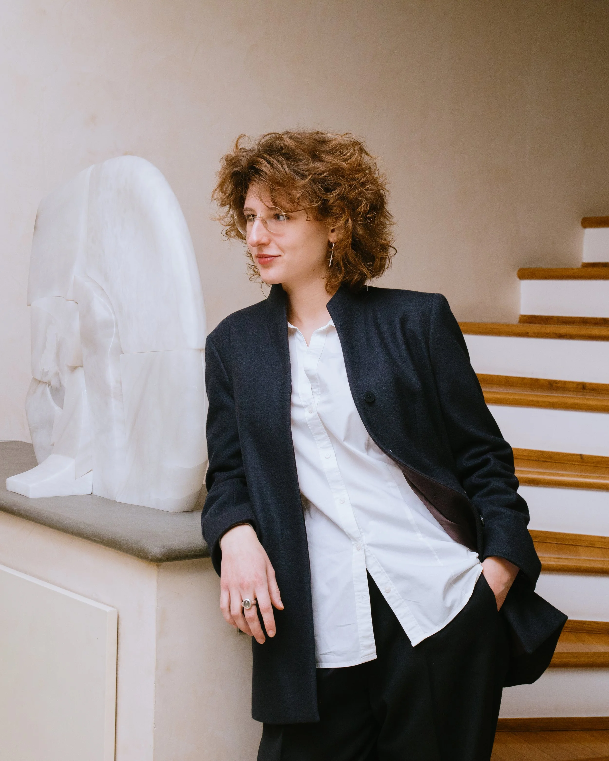 Person with curly hair, glasses, earrings, wearing a dark blazer and white shirt, leaning against a marble sculpture of a horse head, standing on a staircase with wooden steps.