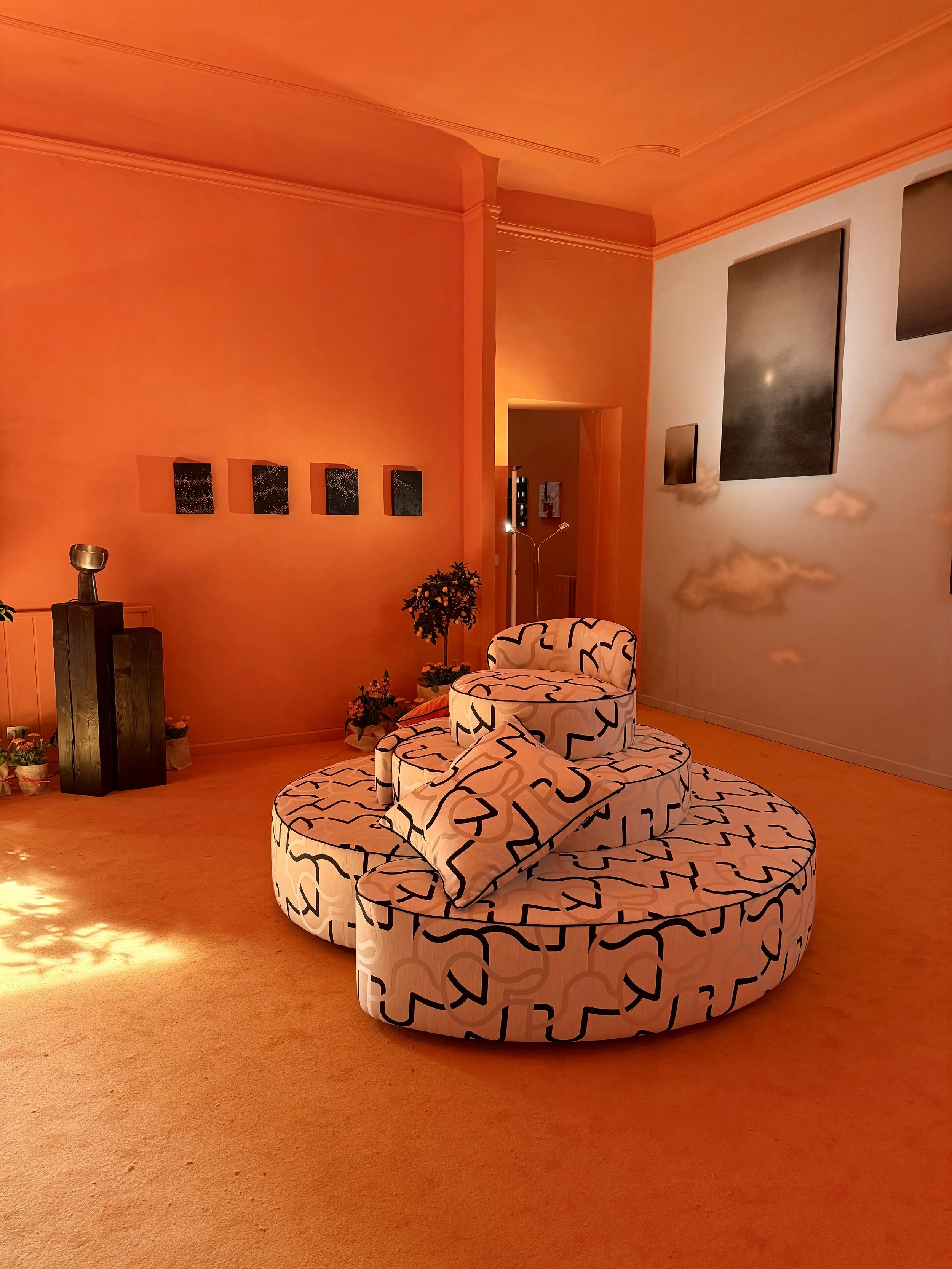 A cozy living room with an orange theme, featuring a round, patterned sofa, a patterned chair, potted plants, wall-mounted decorations, and a flat-screen TV, illuminated by warm lighting.