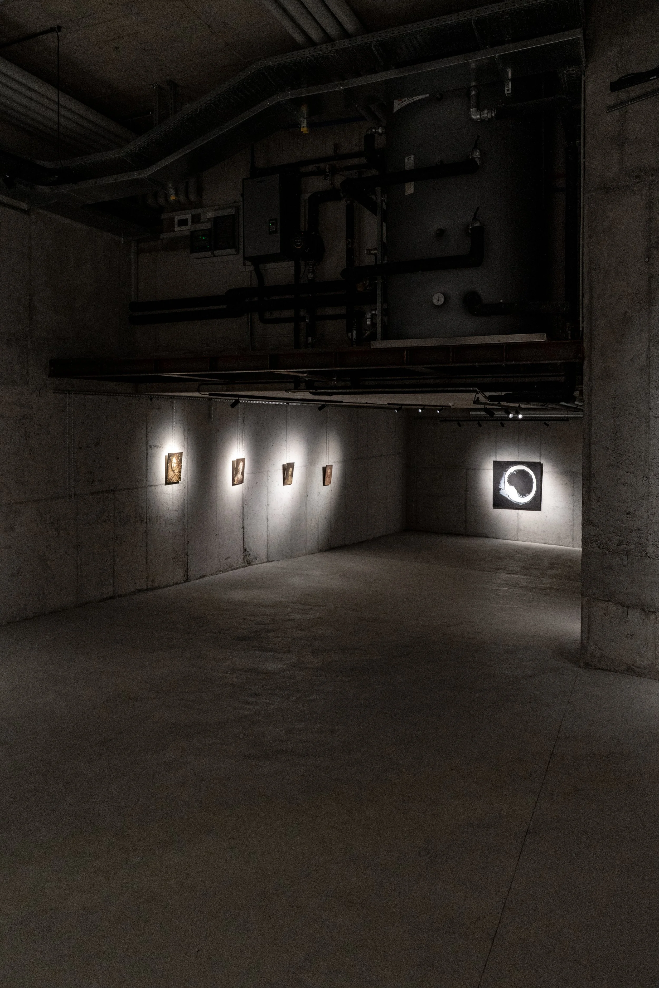 Dimly lit art gallery with a concrete wall and ceiling, displaying small framed artworks along the wall and a illuminated circular artwork at the end of the room.