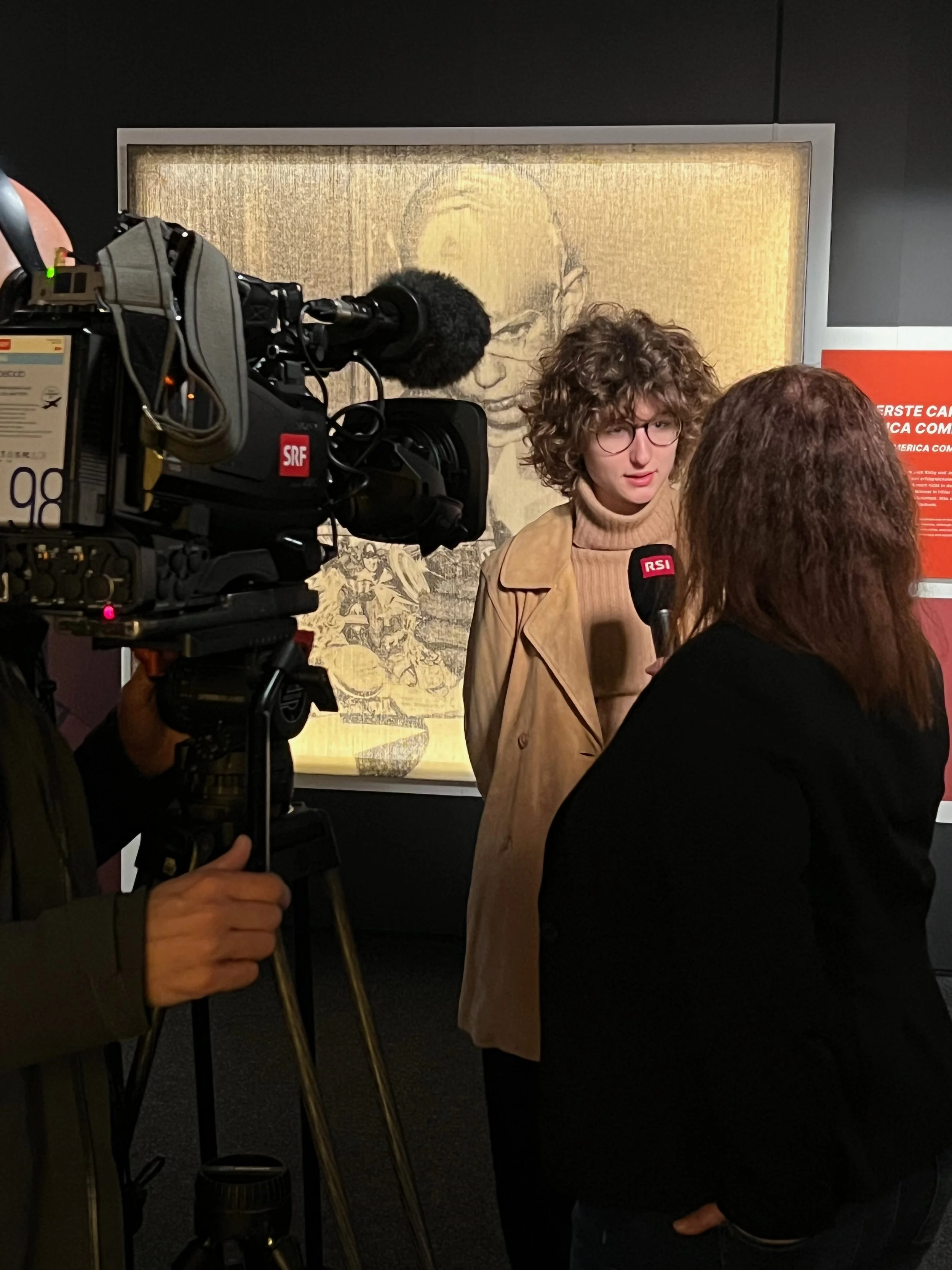 A woman with curly hair and glasses is being interviewed by a person with brown hair at an art exhibit. A camera and microphone labeled 'RSI' are visible, and artwork hangs on the wall behind them.