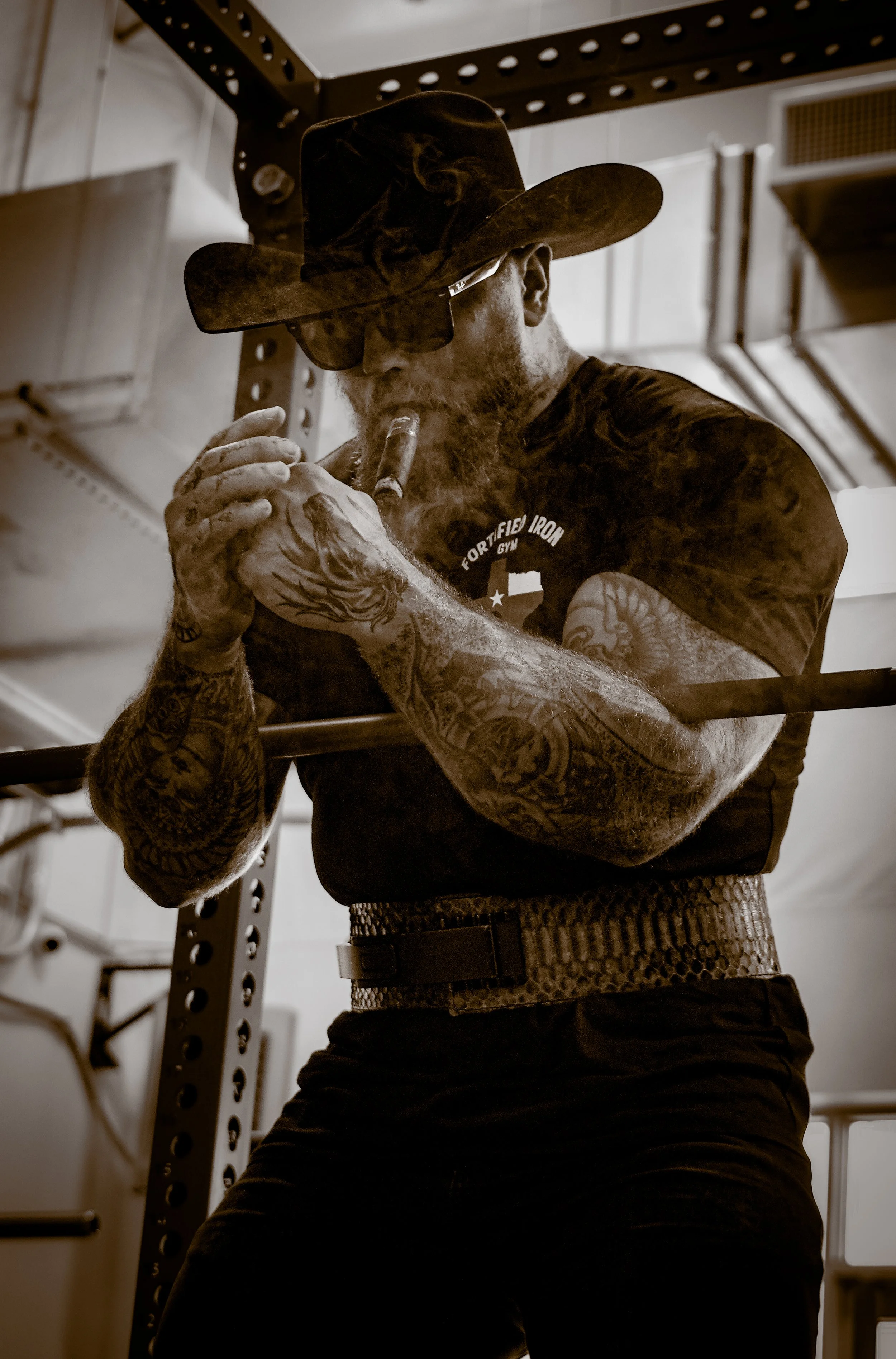 A man with a beard and tattoos on his arms, wearing a cap and sunglasses, is smoking a cigar in a garage or industrial setting.