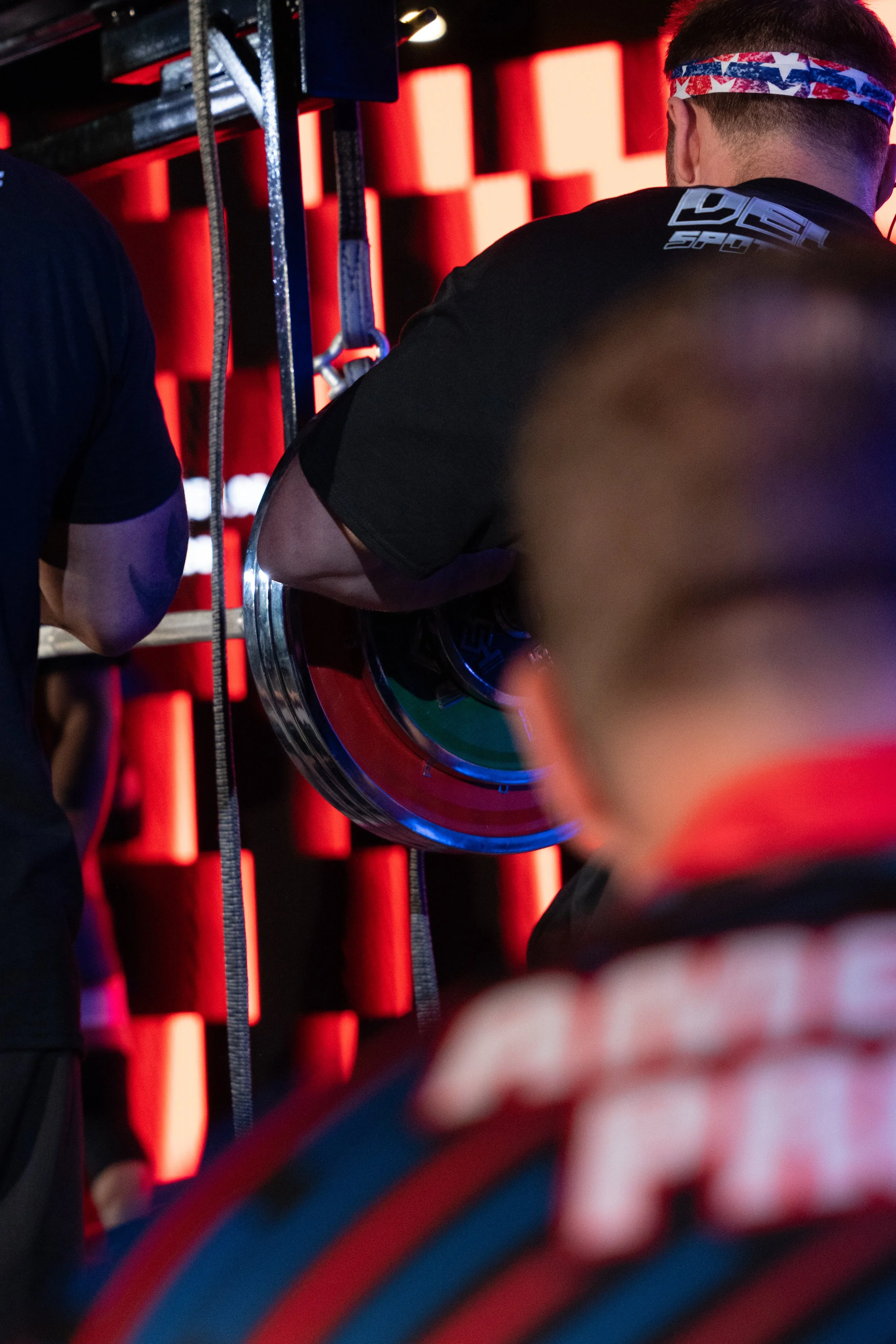 A person with short hair wearing a patriotic headband and a black shirt labeled 'LUSH' on the back, preparing for a fitness activity with a colorful weight plate, in a gym with red padded walls and nearby exercise equipment.