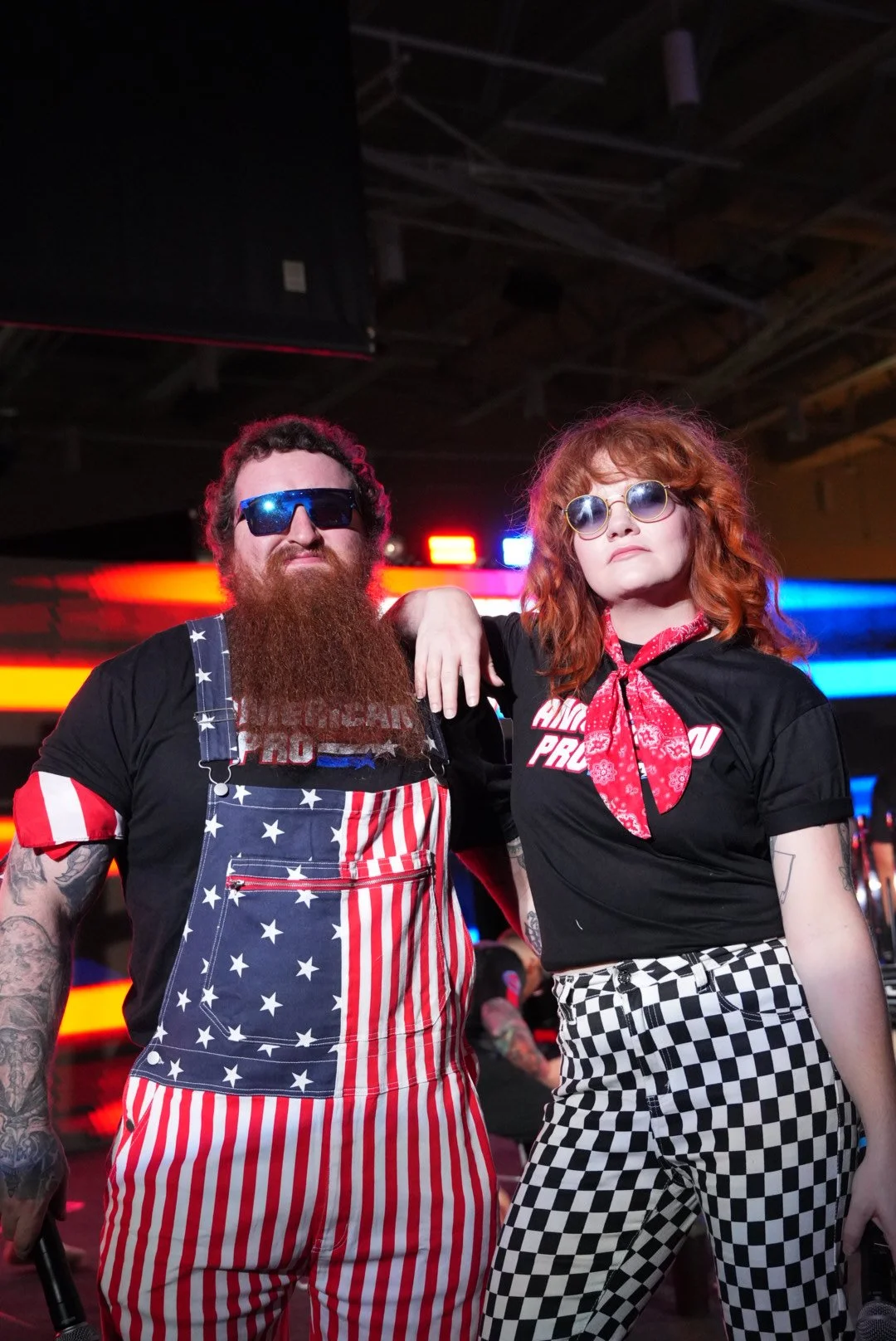 Two people dressed in punk rock style clothing, standing in front of neon lights; the man has a beard, tattoos, sunglasses, and wears an American flag themed apron; the woman has curly red hair, sunglasses, a red bandana, a black T-shirt, and checker