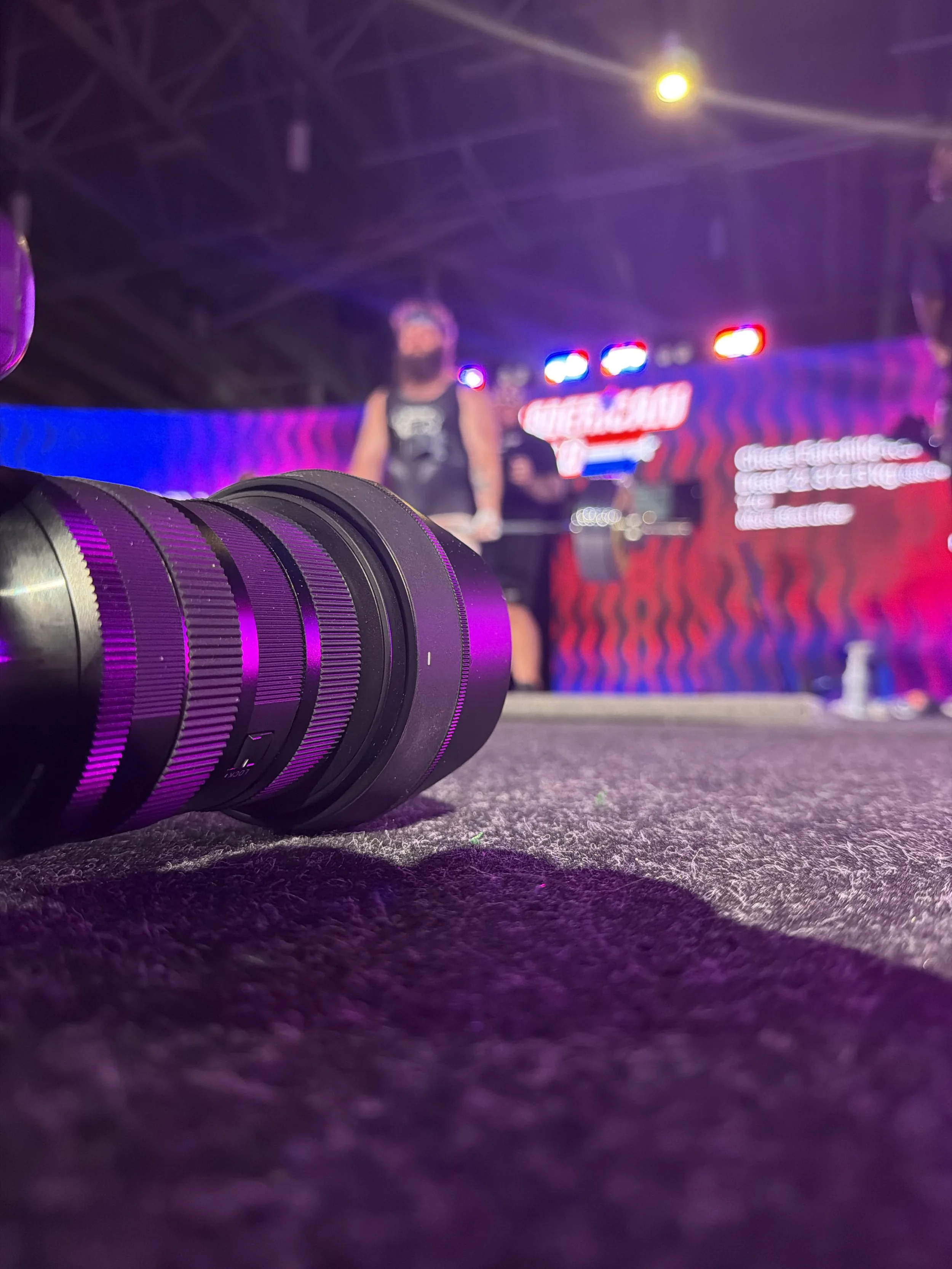 Close-up of a camera lens on a dark, textured surface with stage lighting and blurry LED screens in the background at a concert or event.