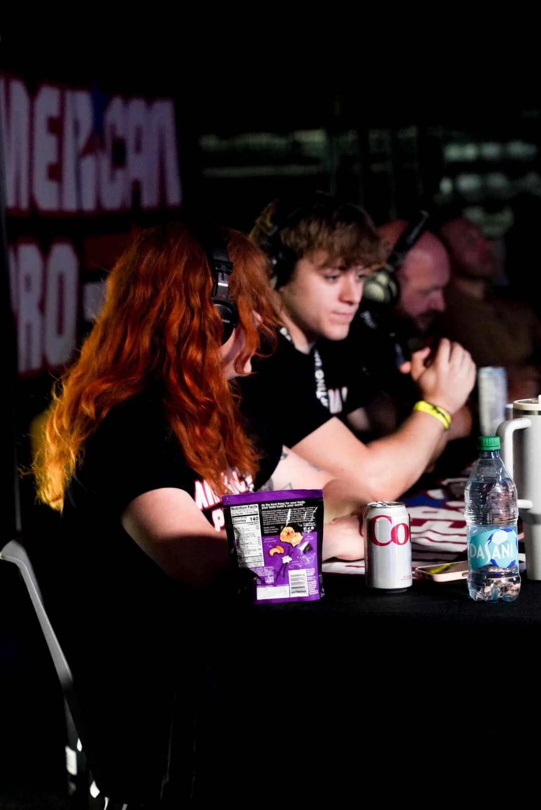 Three individuals sitting at a table with headphones on, focusing on something in front of them; drinks and snacks on the table.