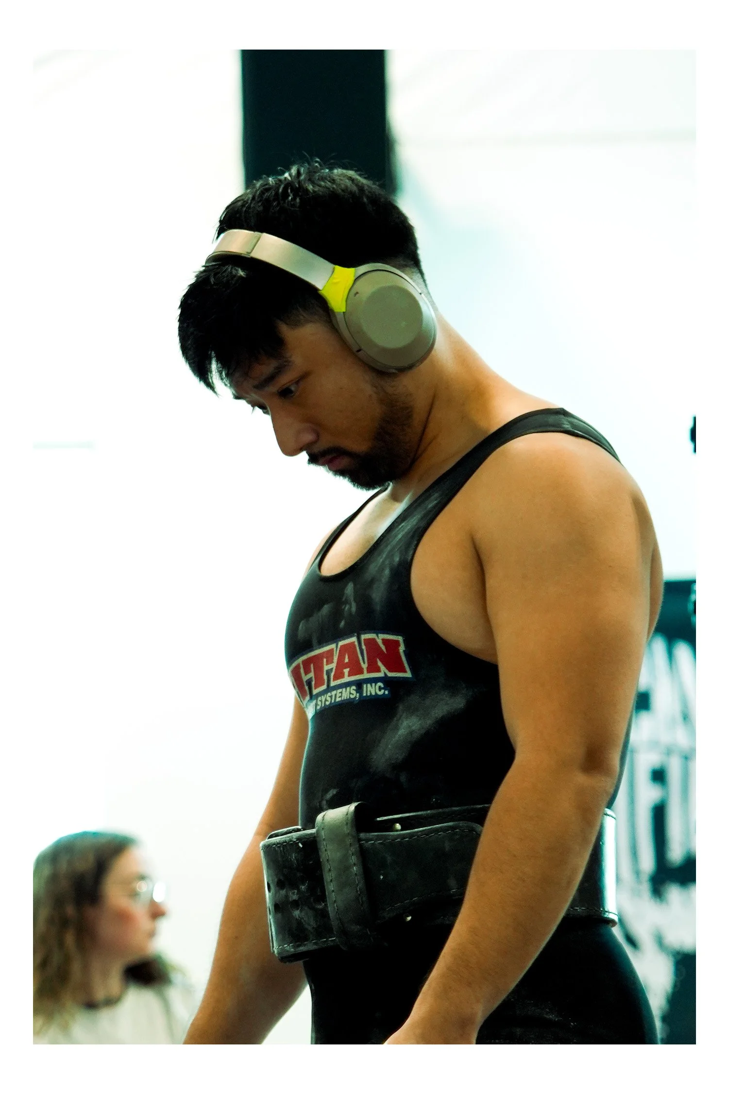 A young man with dark hair and facial hair wearing large over-ear headphones and a black tank top with a red and white logo, standing with his head bowed down. There is a woman with glasses and light-colored hair blurred in the background.