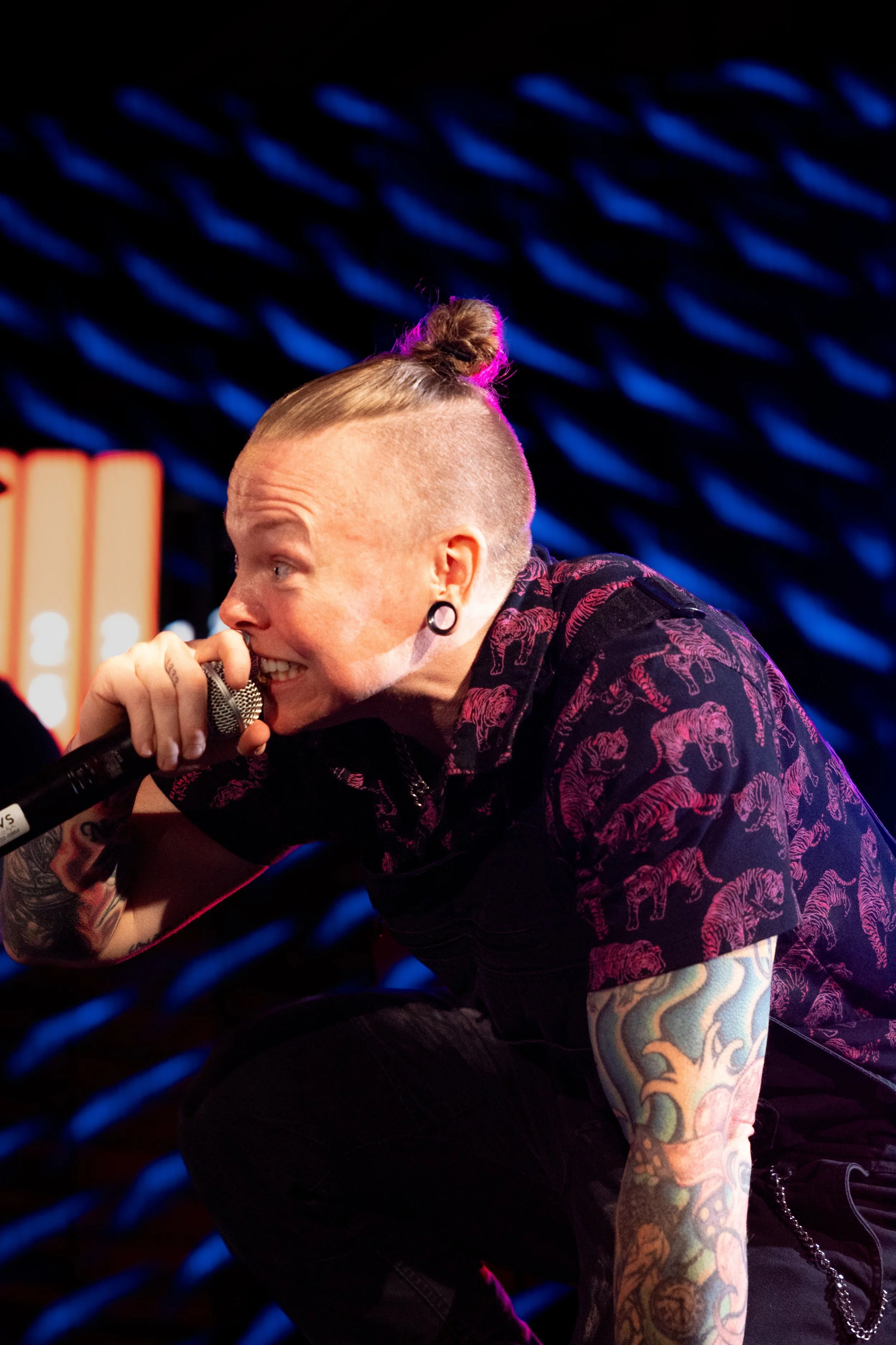 A person with shaved sides and an updo hairstyle, wearing a black shirt with pink tiger print, holding a microphone, with tattoos on their arm, in front of a background with blue diagonal lines.