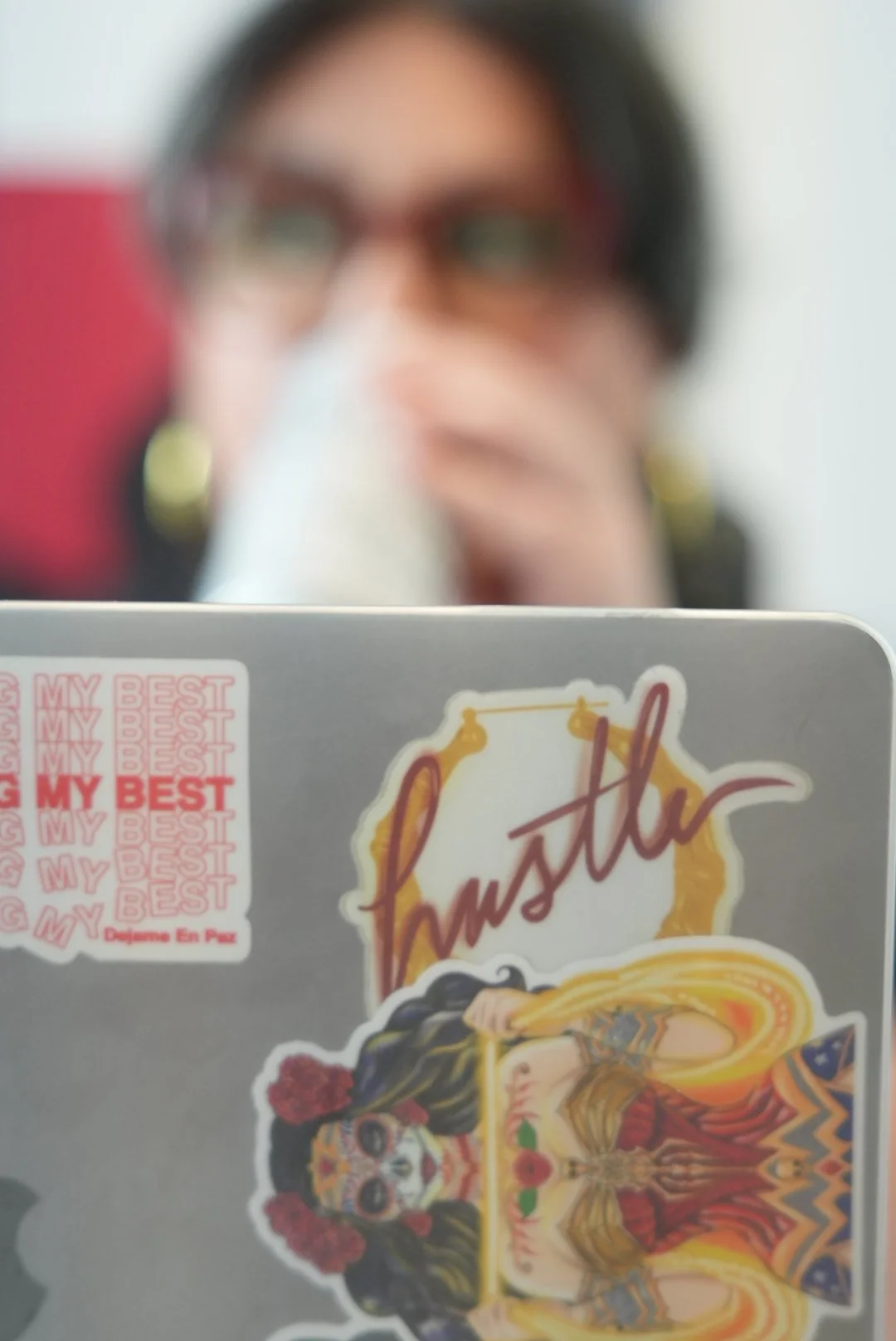 A person holds a laptop decorated with colorful stickers, including one that says "My Best" repeated in pink text, and a large sticker with a woman in a gold dress and tattoos. The person is out of focus with glasses and earrings, and the background 