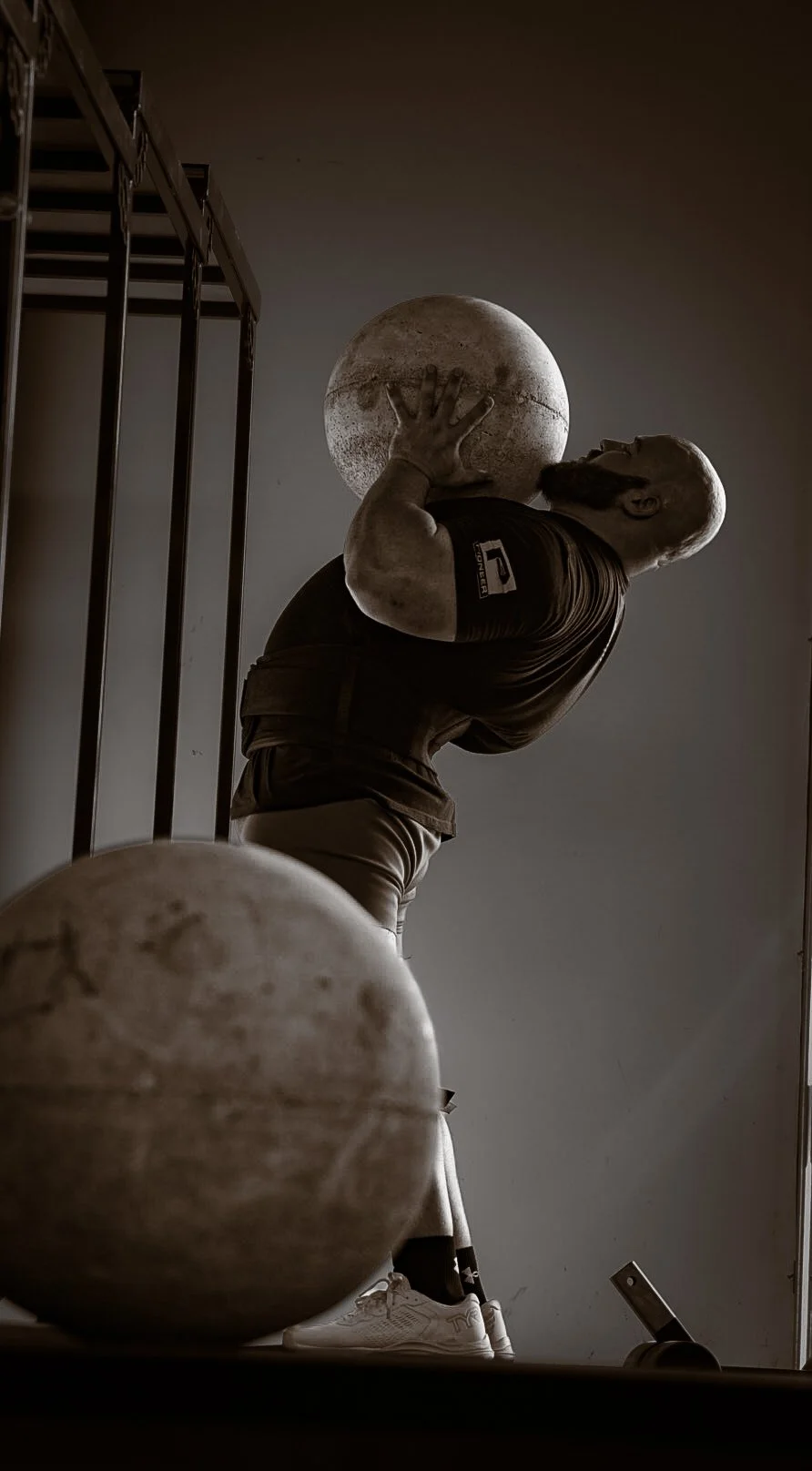 A man with a muscular build, a beard, and athletic clothing is holding a textured medicine ball close to his chest during a workout. The photo is taken from a low angle, emphasizing the effort of the lift.