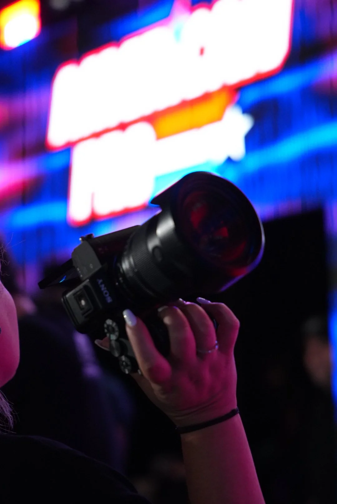 Close-up of a person holding a camera, with a blurred colorful neon sign in the background.