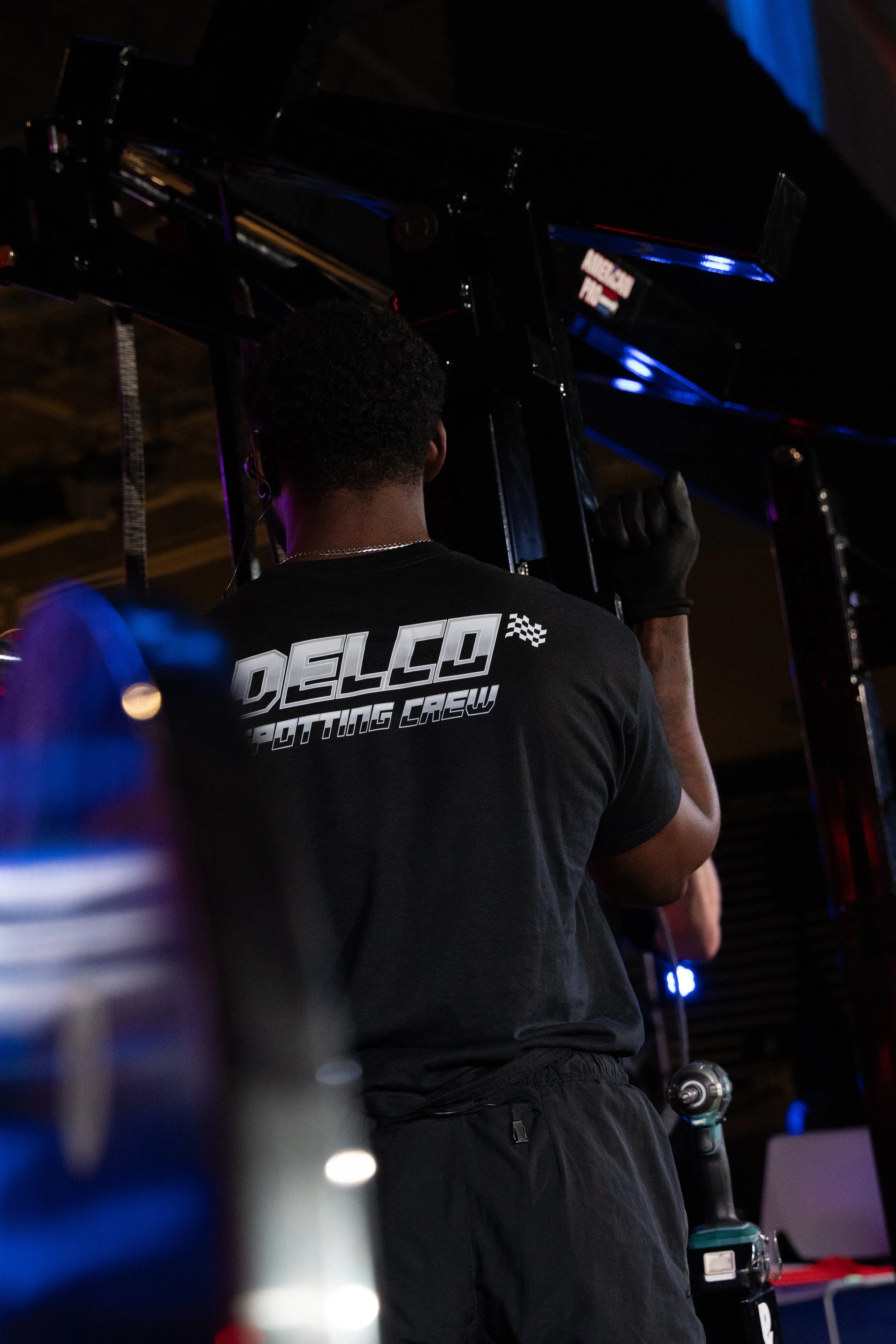 A person wearing a black shirt with 'DELCO. POTTING CREW' printed on the back, working with a tool in a dimly lit environment, possibly a workshop or garage.