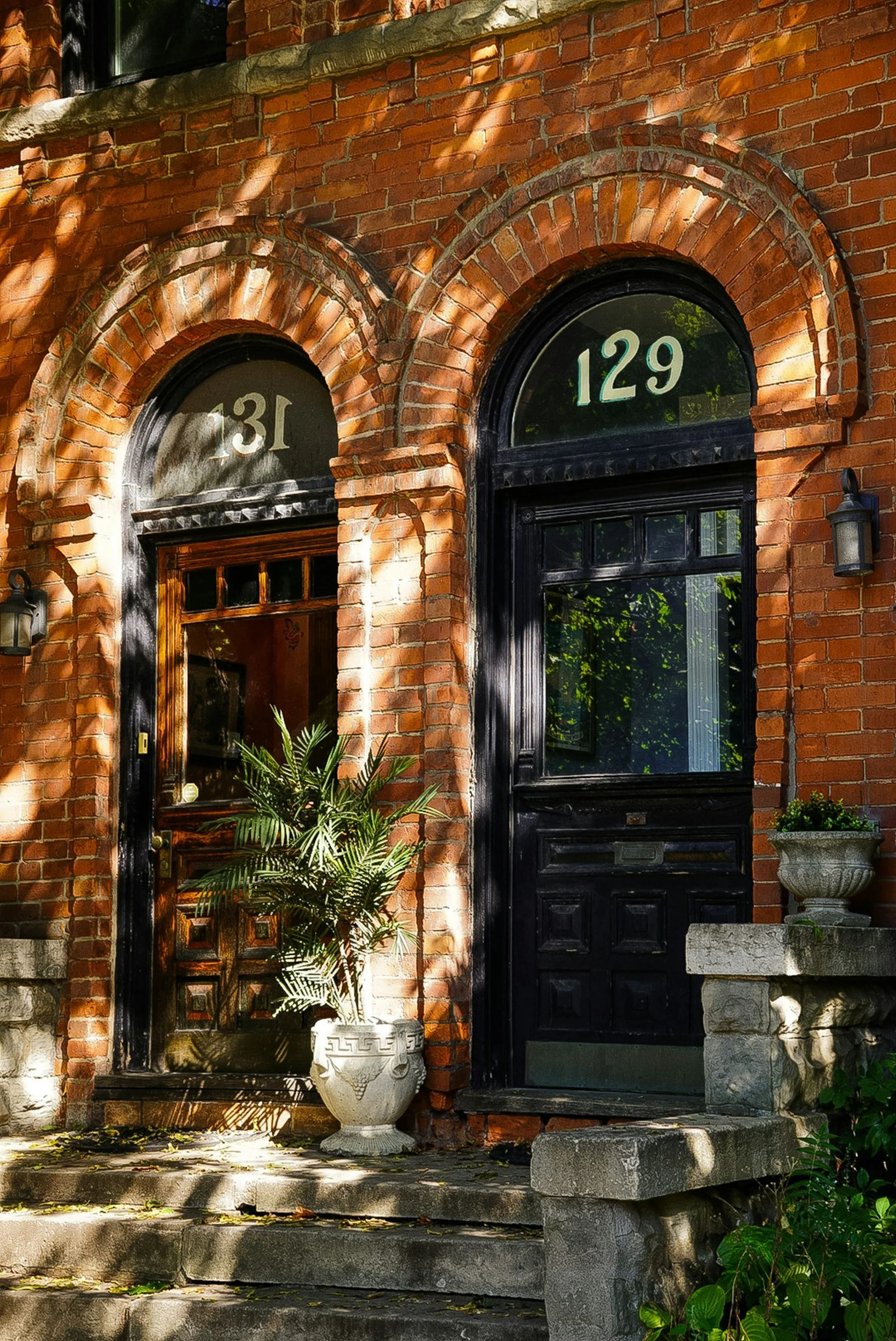 The image shows the front entrance to a brick house with two arched windows and a wooden door. The house has house numbers 31 and 129 above the windows. There are plants in white and stone planters near the entrance, and a stone step leading up to the porch, with dappled sunlight and shadows.