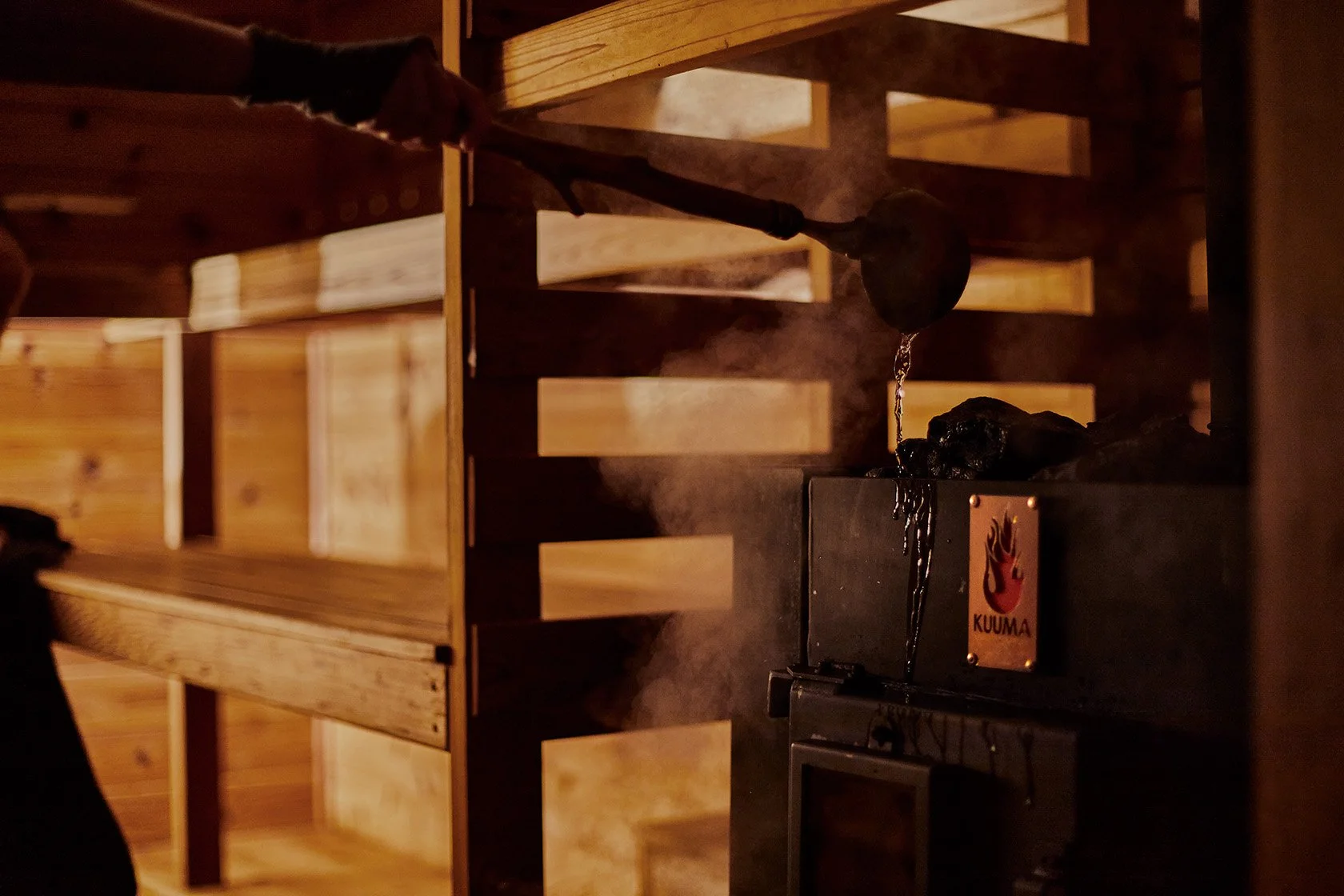 Dark wood-fired sauna interior with a steaming Kuuma black metal stove and wooden benches.