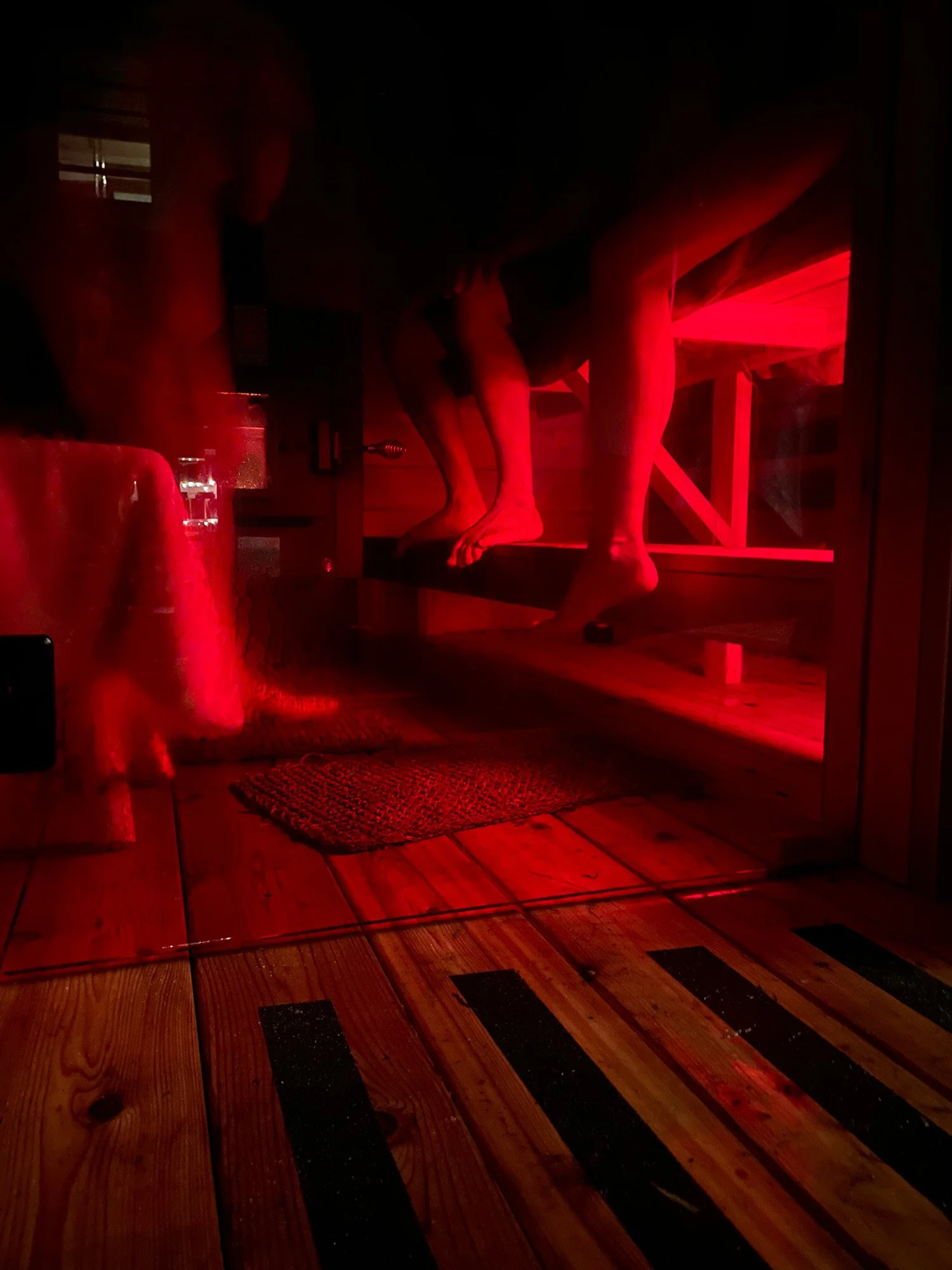 Dimly lit red room with a person’s bare feet resting on a wooden bench, a drum, and wood plank flooring in a wooden sauna