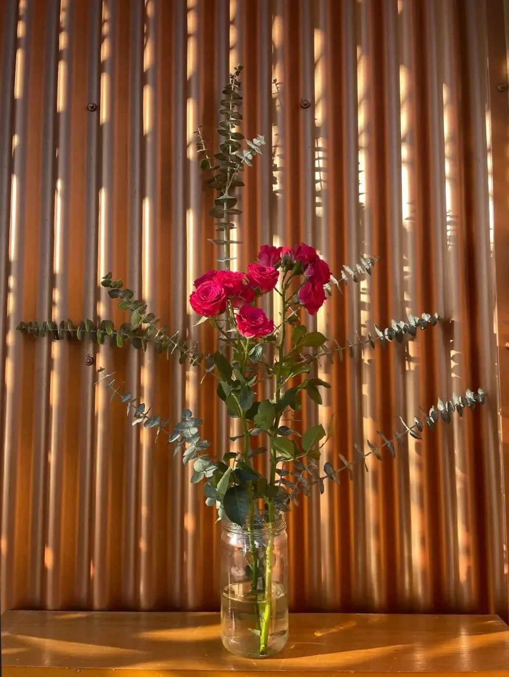 Bouquet of pink roses and eucalyptus branches in a glass jar against a wood-paneled wall