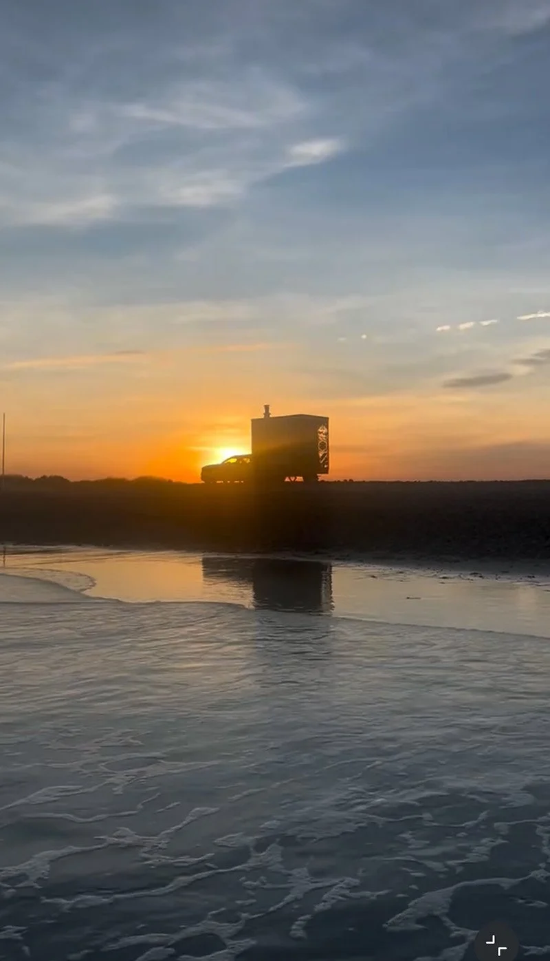 Silhouetted truck parked on a shoreline at sunset, reflected in shallow water.