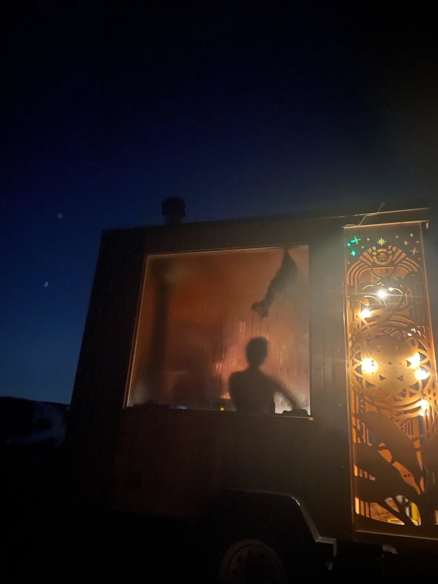 Nighttime view of a mobile sauna trailer with glowing patterned panels and silhouettes visible through a service window