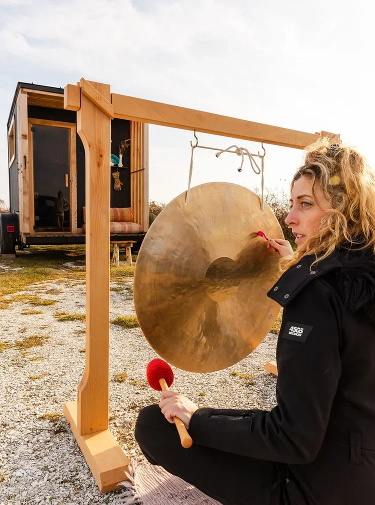 Woman striking a large hanging gong with a mallet outdoors beside mobile sauna
