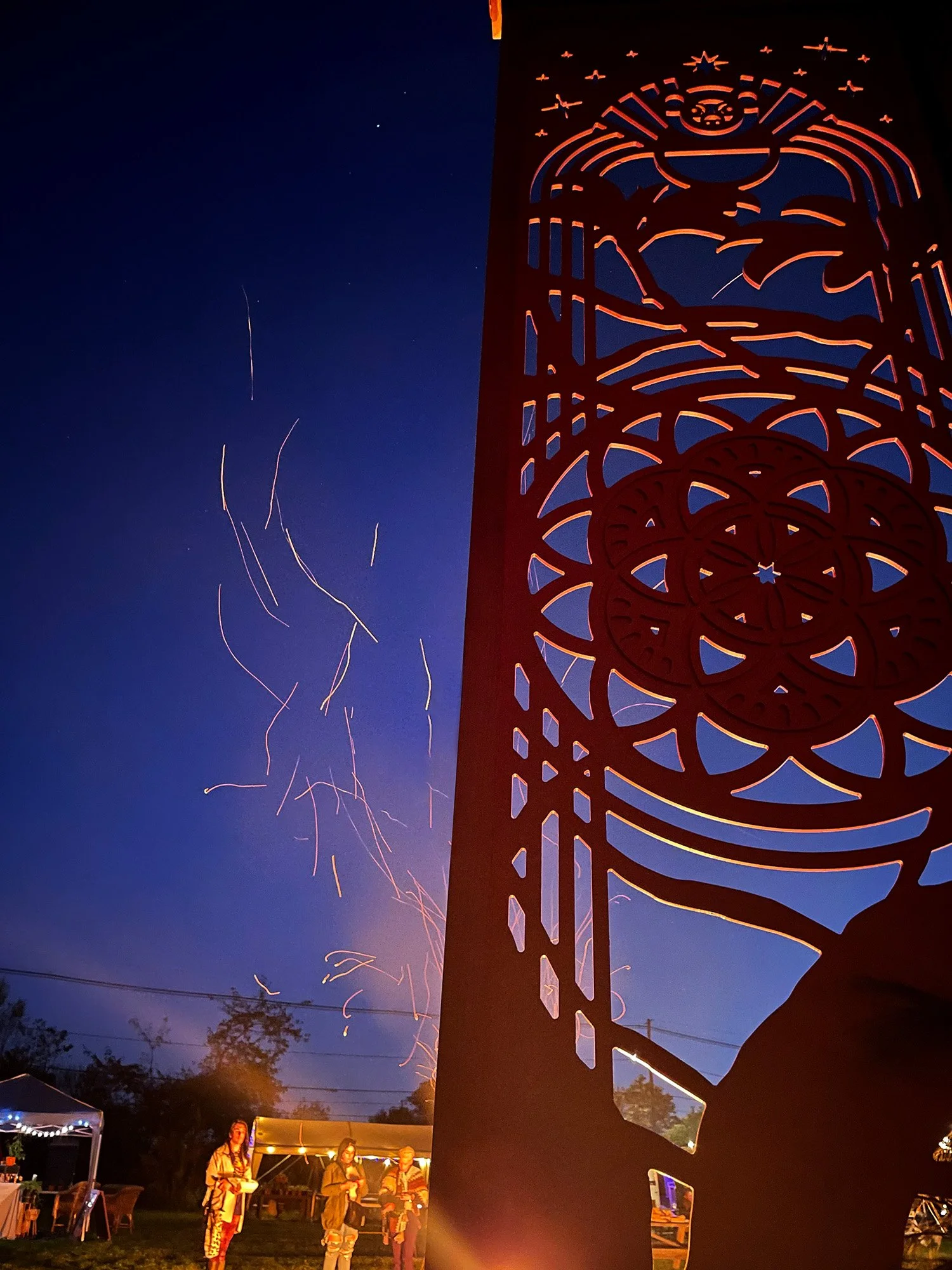 Illuminated decorative metal sculpture with a stylized face and geometric patterns at an outdoor nighttime festival, with fireworks and people in the background.