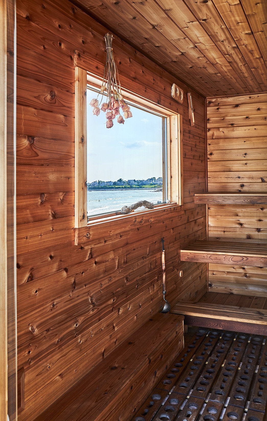 Rustic wooden beach sauna interior with benches, dried flowers, a paddle, and ocean view through the window