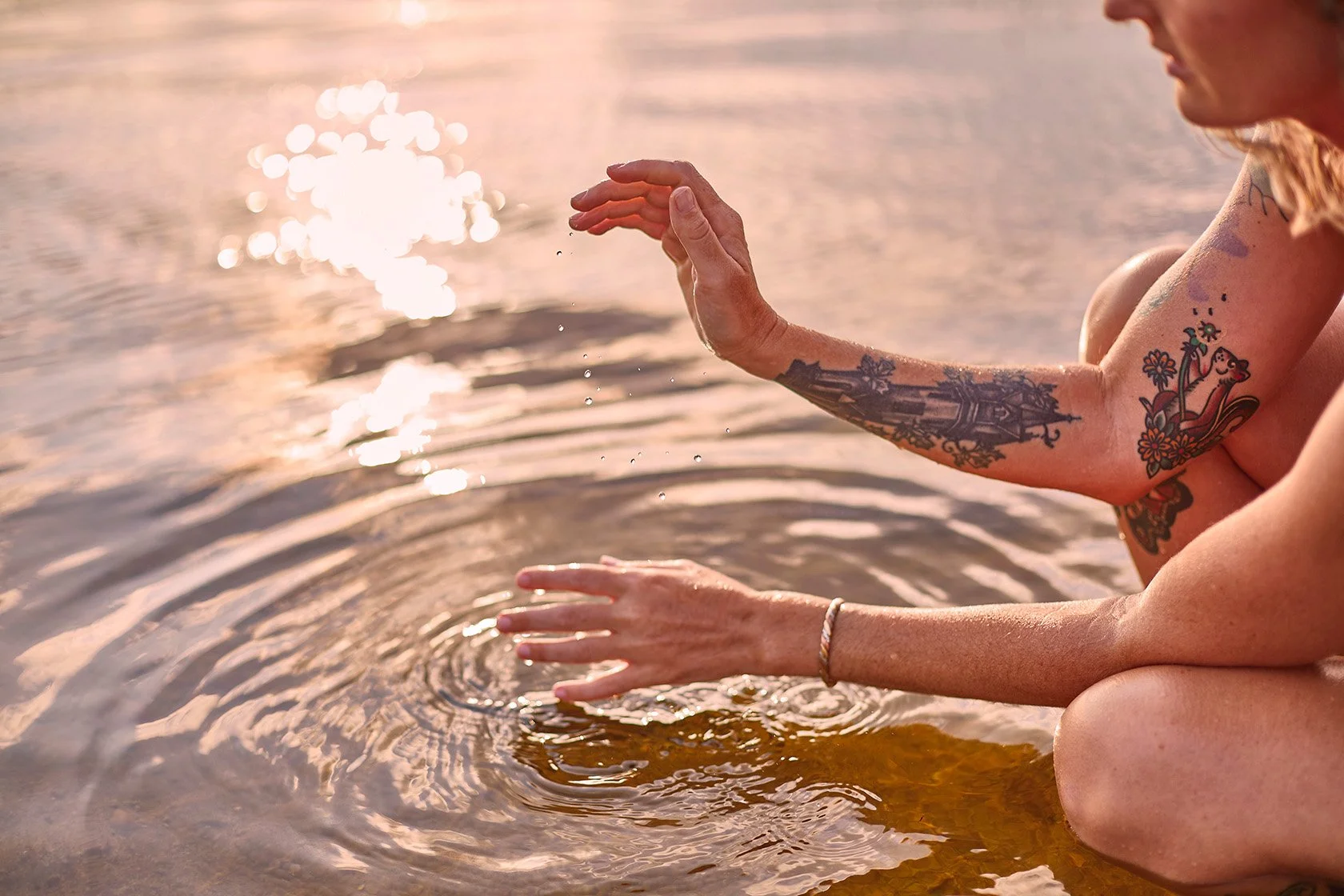 Tattooed woman touching sunlit water at sunset