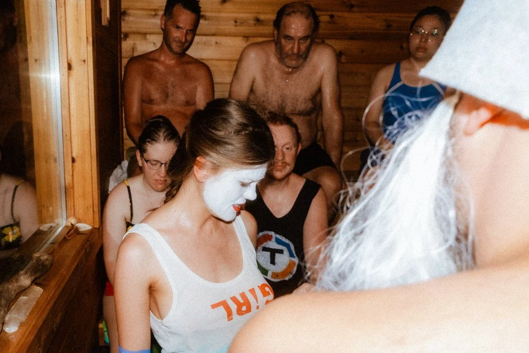 Group of people gathered inside a wood-paneled sauna, including a woman wearing white face paint and a white tank top.