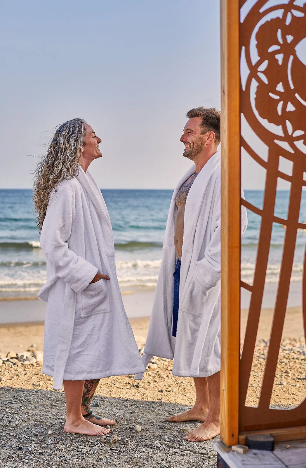 Smiling couple in white bathrobes standing barefoot on a beach beside the ocean