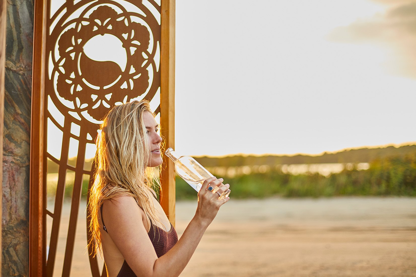 Blonde woman drinking from a glass bottle beside a decorative window overlooking a sunlit lake