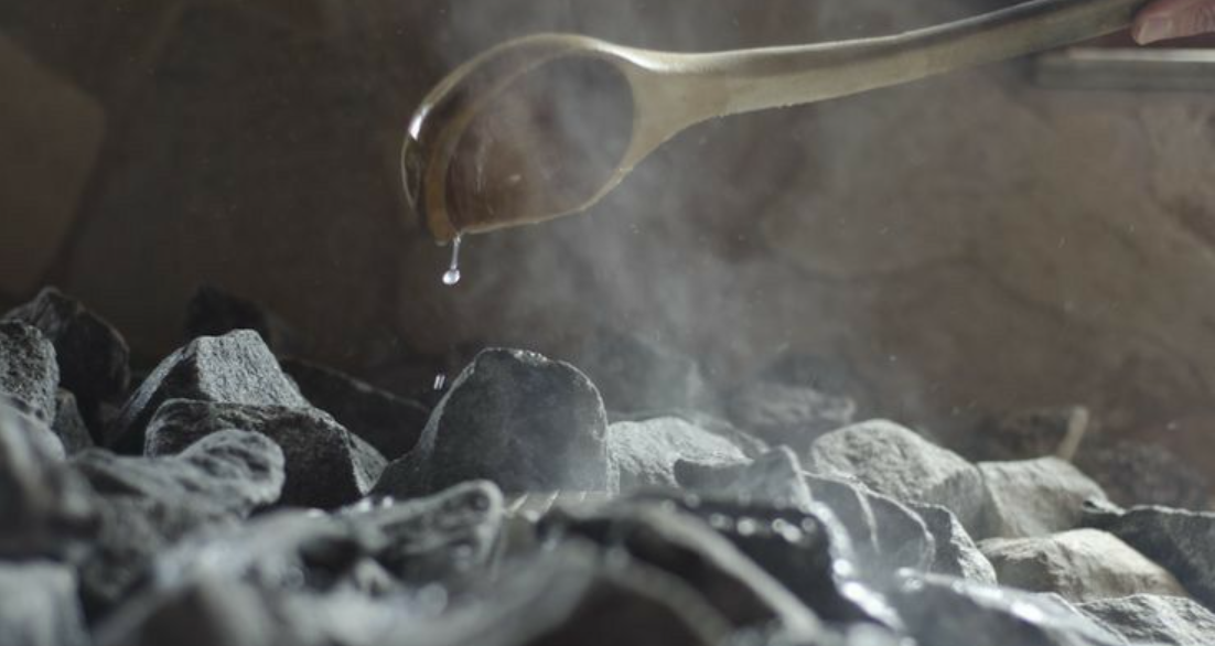 Metal spoon holding a droplet of water above hot sauna stones with rising steam