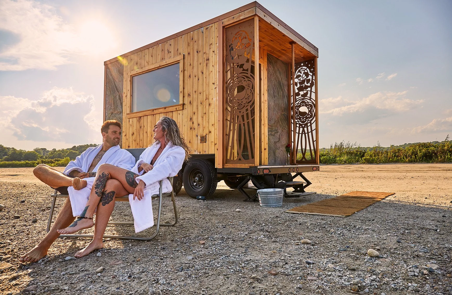 Couple in white robes relaxing beside a mobile wooden sauna trailer at a beach at sunset