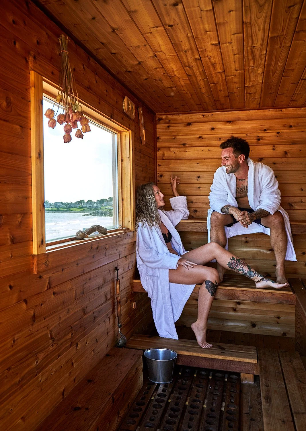 Couple in white robes relaxing inside a rustic wooden mobile sauna with a ocean view through the window