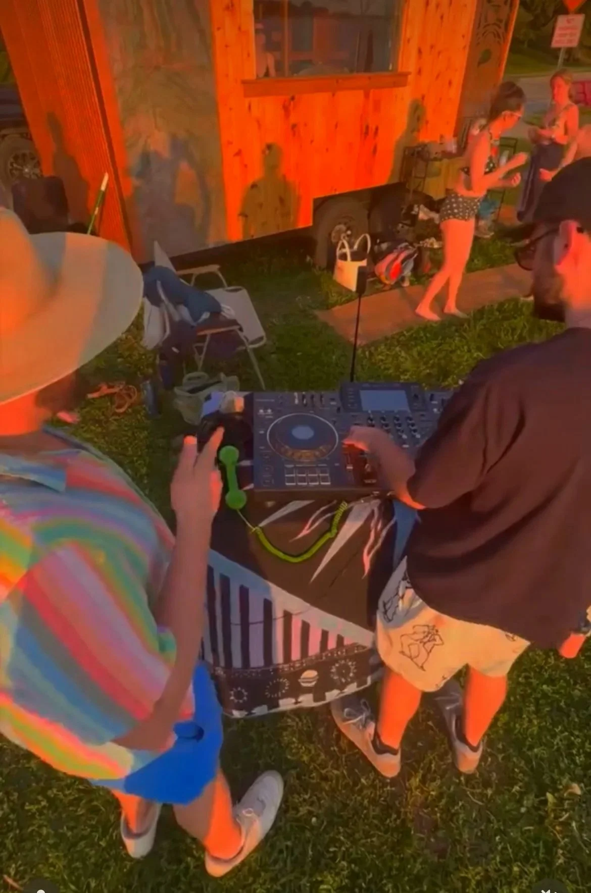People dancing outdoors beside a wooden mobile sauna while a DJ operates a controller at a sunset party.