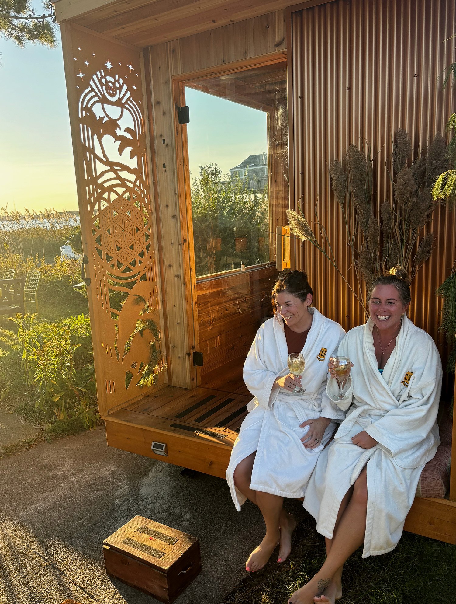 Two smiling women in white robes holding wine glasses beside a decorated wooden outdoor sauna overlooking a sunny waterfront