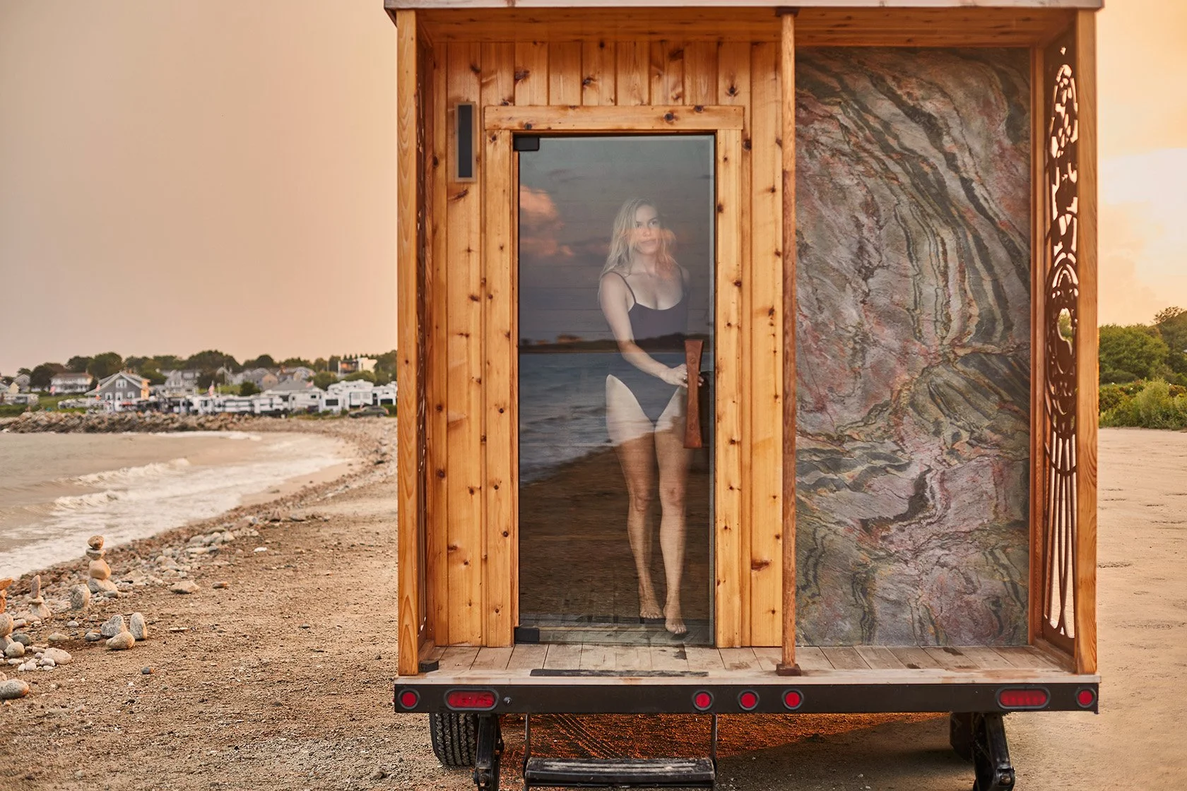 Wooden beach sauna trailer with a woman visible through the glass door on a sandy shoreline at sunset