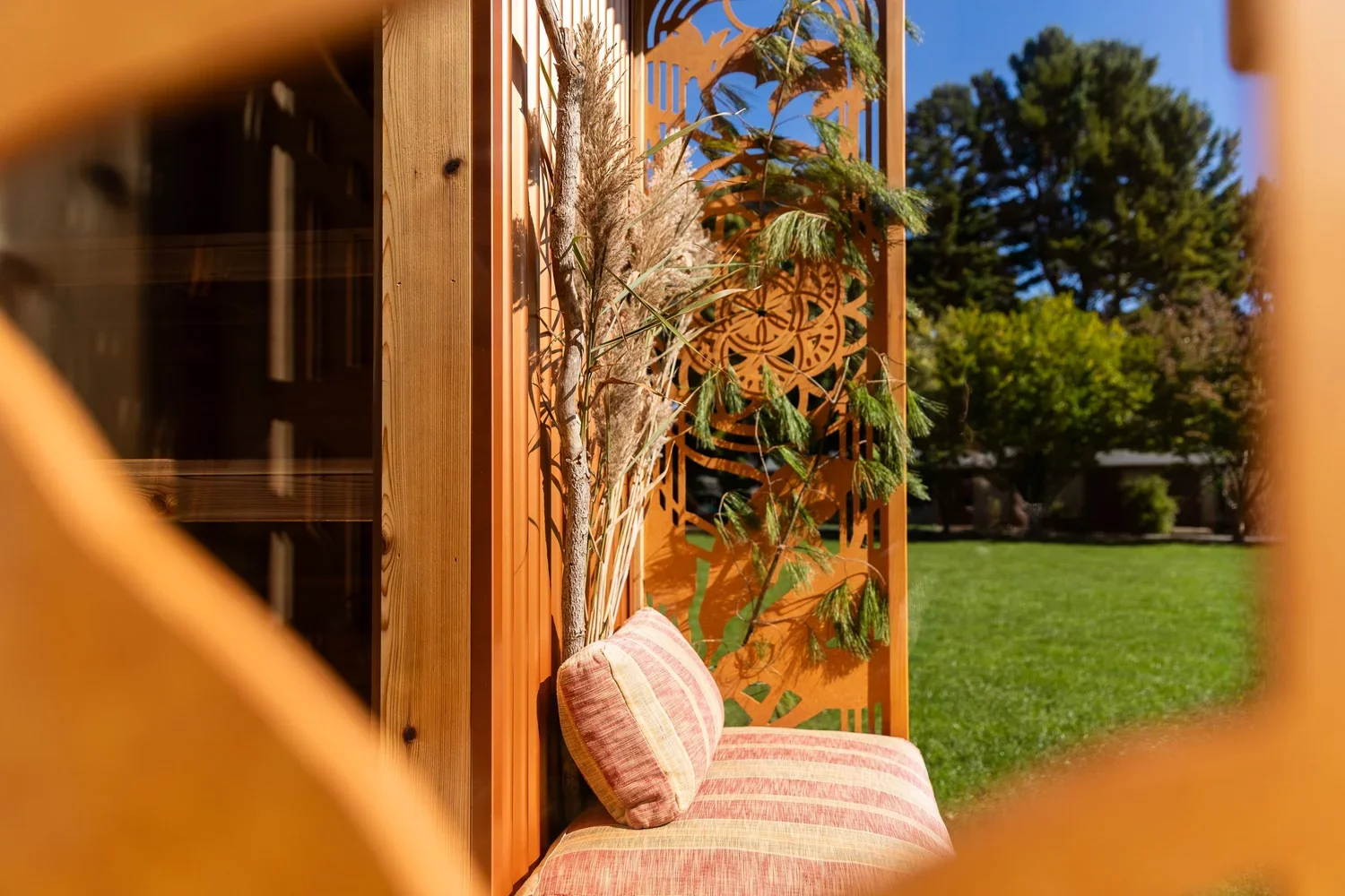 Striped cushioned outdoor lounge chair on a mobile sauna beside a carved wooden privacy screen and potted plant in a sunny garden