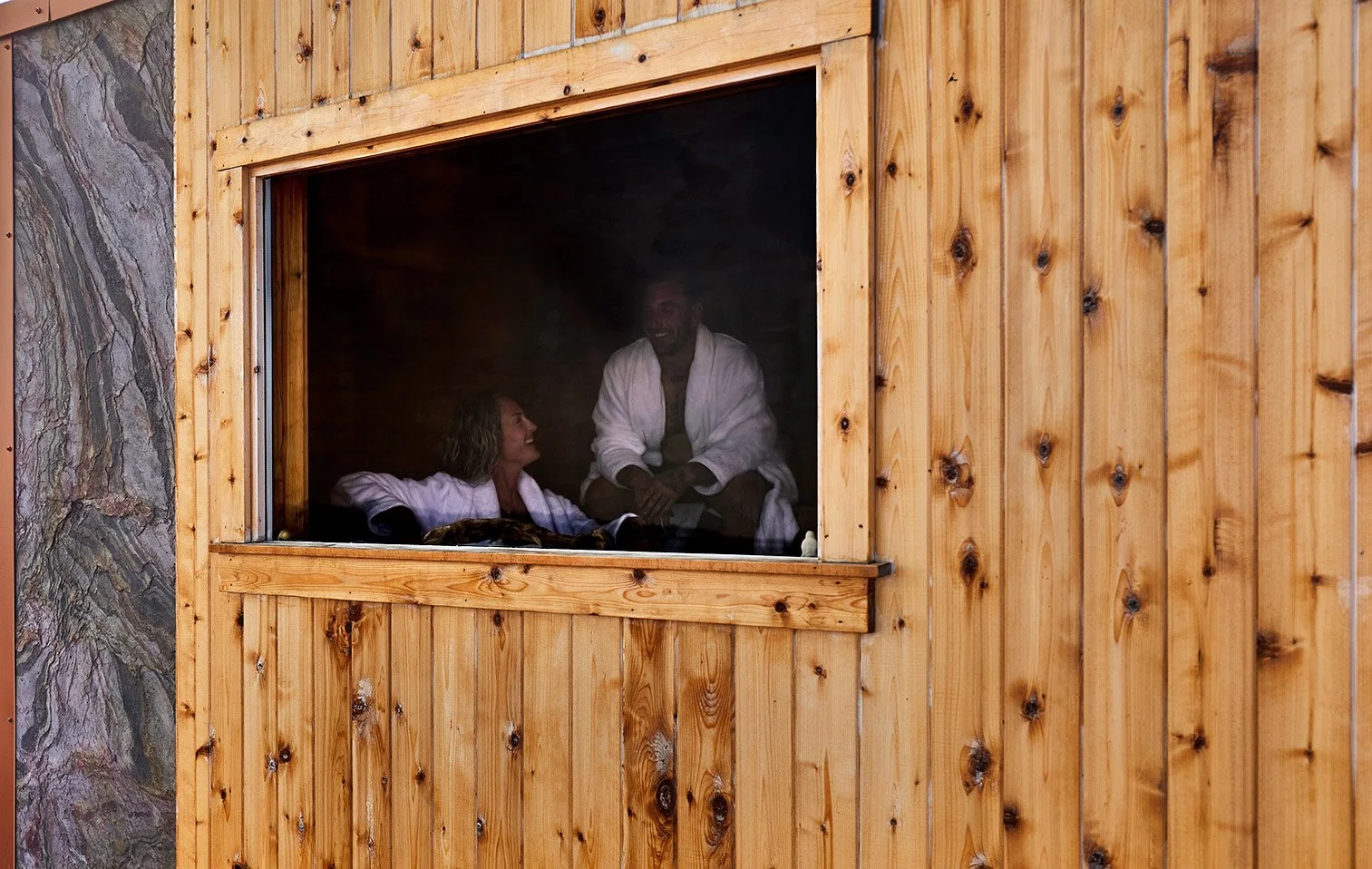 Two people in white robes relaxing inside a wooden sauna, viewed through an open window.