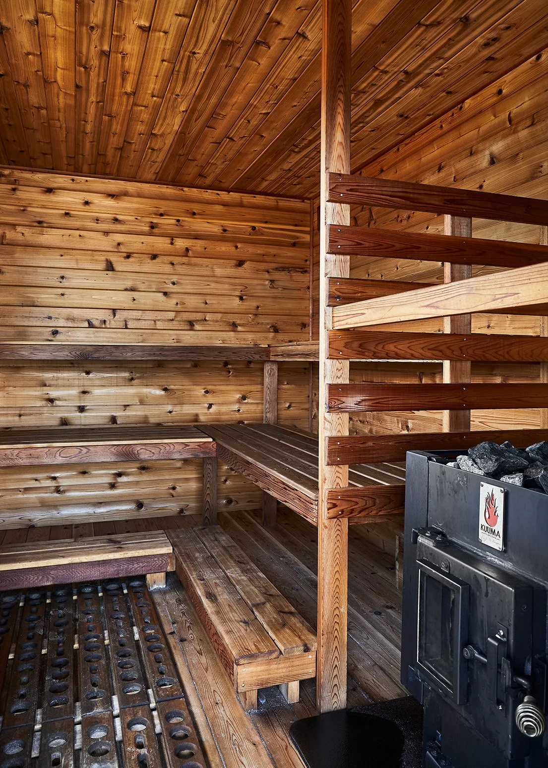 Wood-paneled sauna interior with tiered benches, wooden slat divider, and black Kuuma wood-burning sauna stove filled with stones