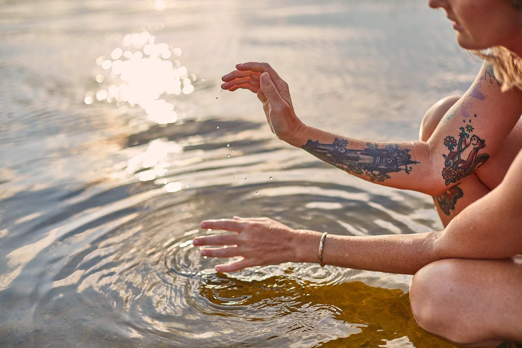 Tattooed woman sitting in sunlit water, gently touching the surface and creating ripples
