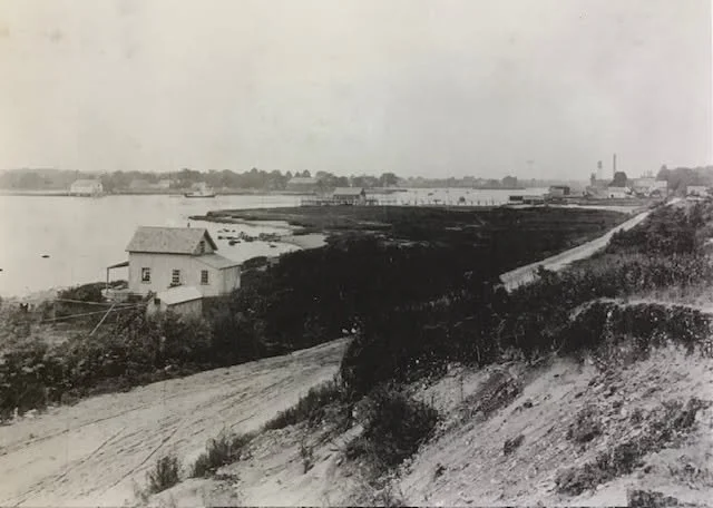 Historic black-and-white photograph of a coastal village with a small house, dirt road, shoreline, docks, and distant buildings.
