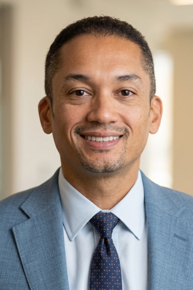 A man in a light blue suit, white shirt, and navy tie with small patterns, smiling indoors with a blurred background.