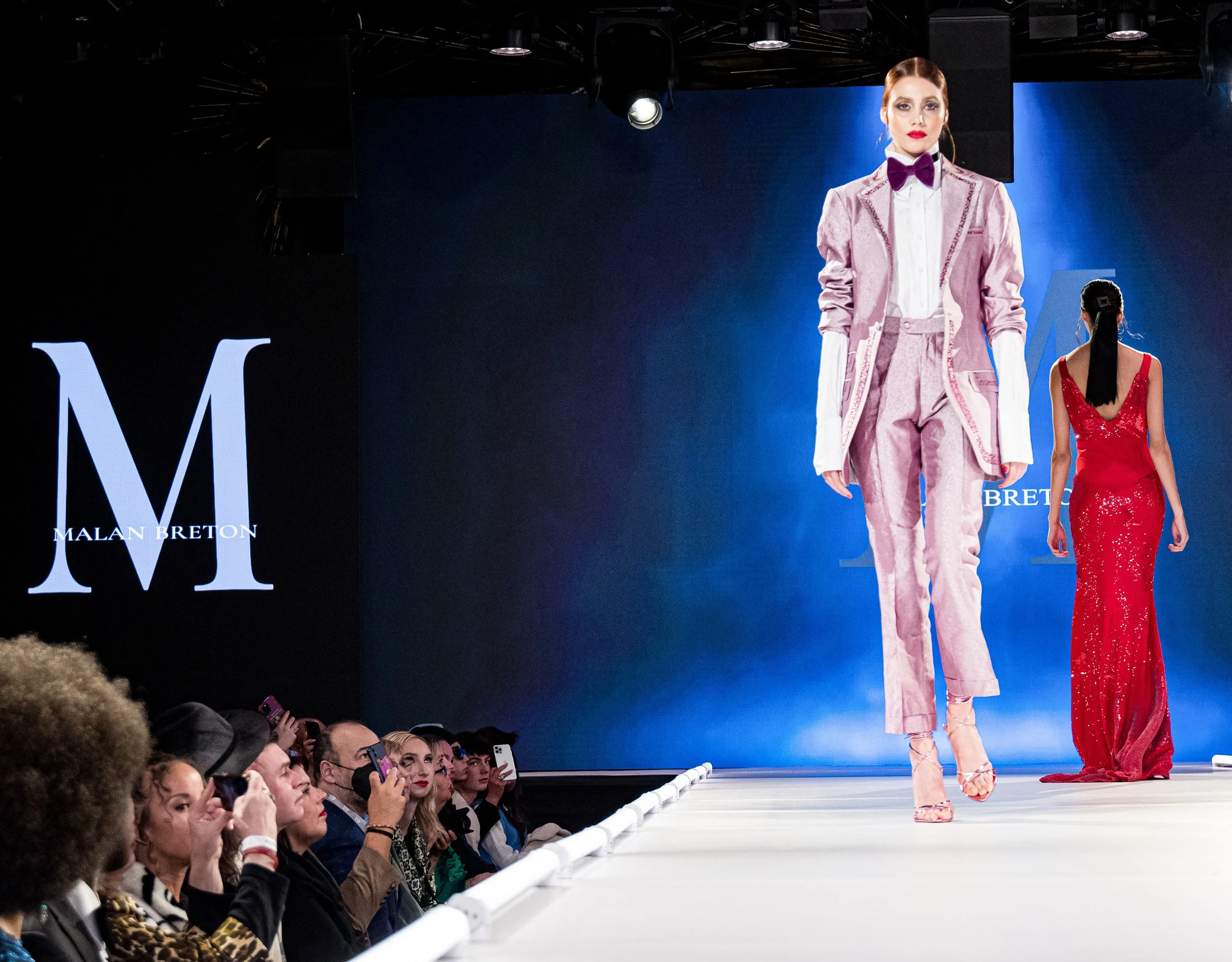 Fashion models walk down the runway during a fashion show, with a large blue screen in the background displaying the name "Malan Breton" and "Bret." The audience watches and takes photos.