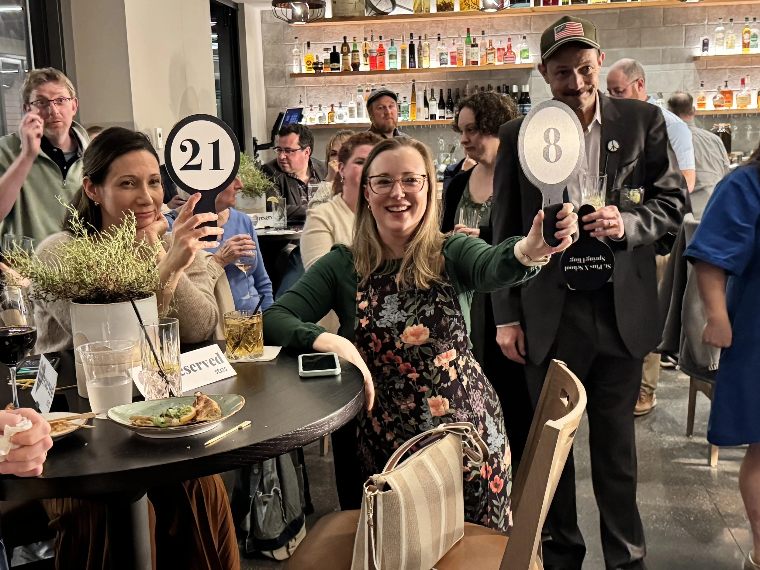 A group of people at a restaurant or party, with some holding signs numbered 21 and 8. They are smiling and enjoying themselves, with drinks and food on the table. The background shows a bar with bottles of alcohol on shelves.