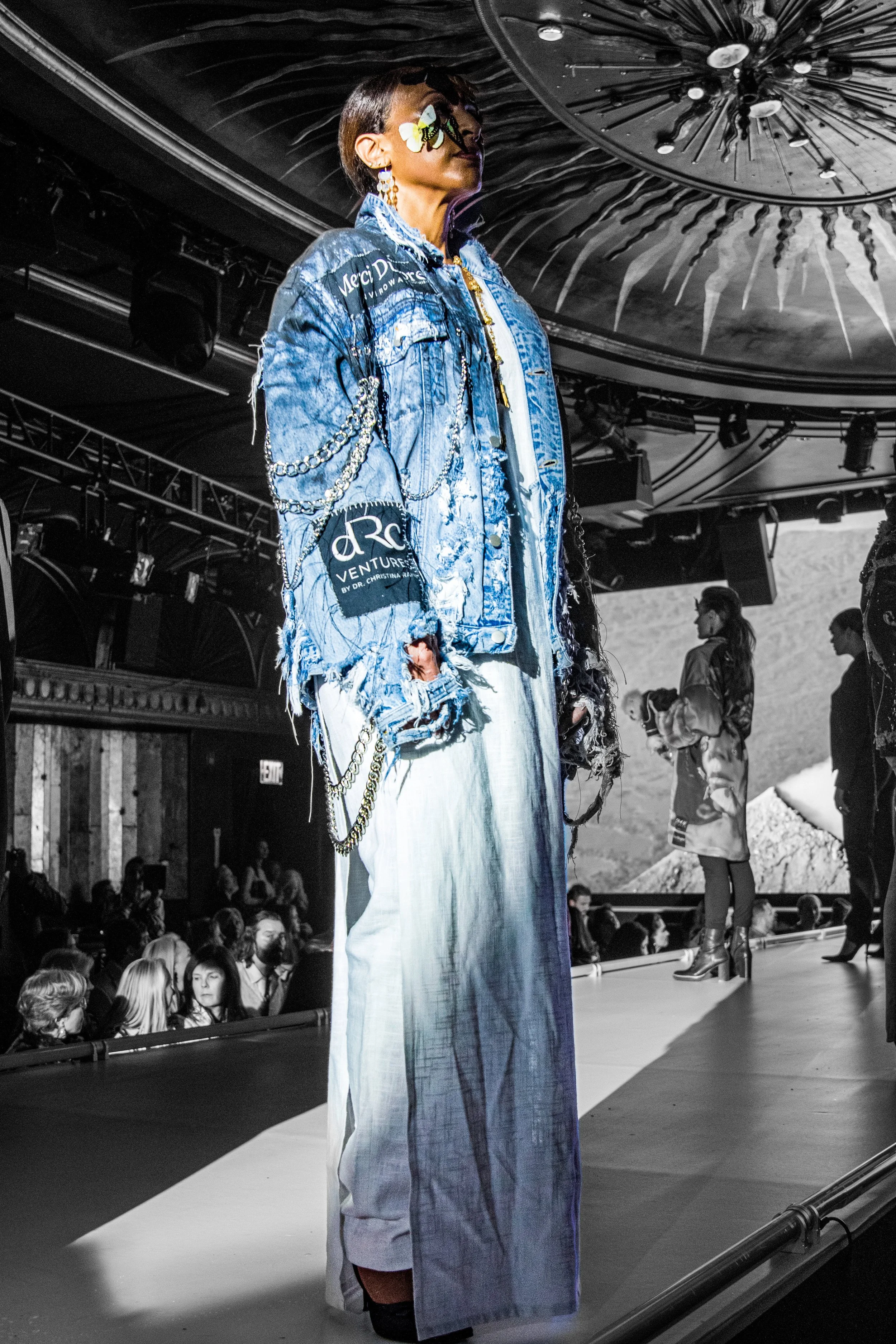 A woman with short hair and sunglasses decorated with flowers on a runway in a fashion show, wearing a distressed denim jacket with patches and chains, and wide-legged jeans.