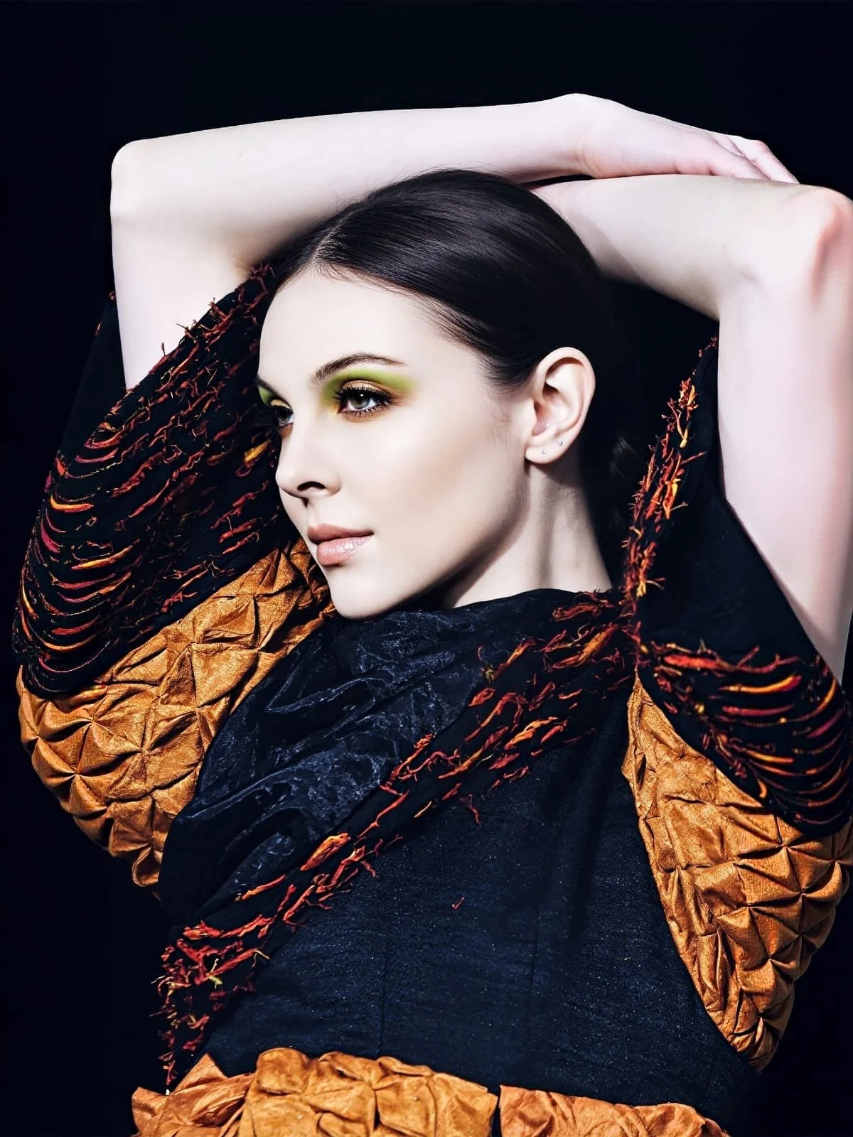A woman with dark hair and striking green eyeshadow poses with her arms raised above her head against a black background, wearing a textured black and orange garment.
