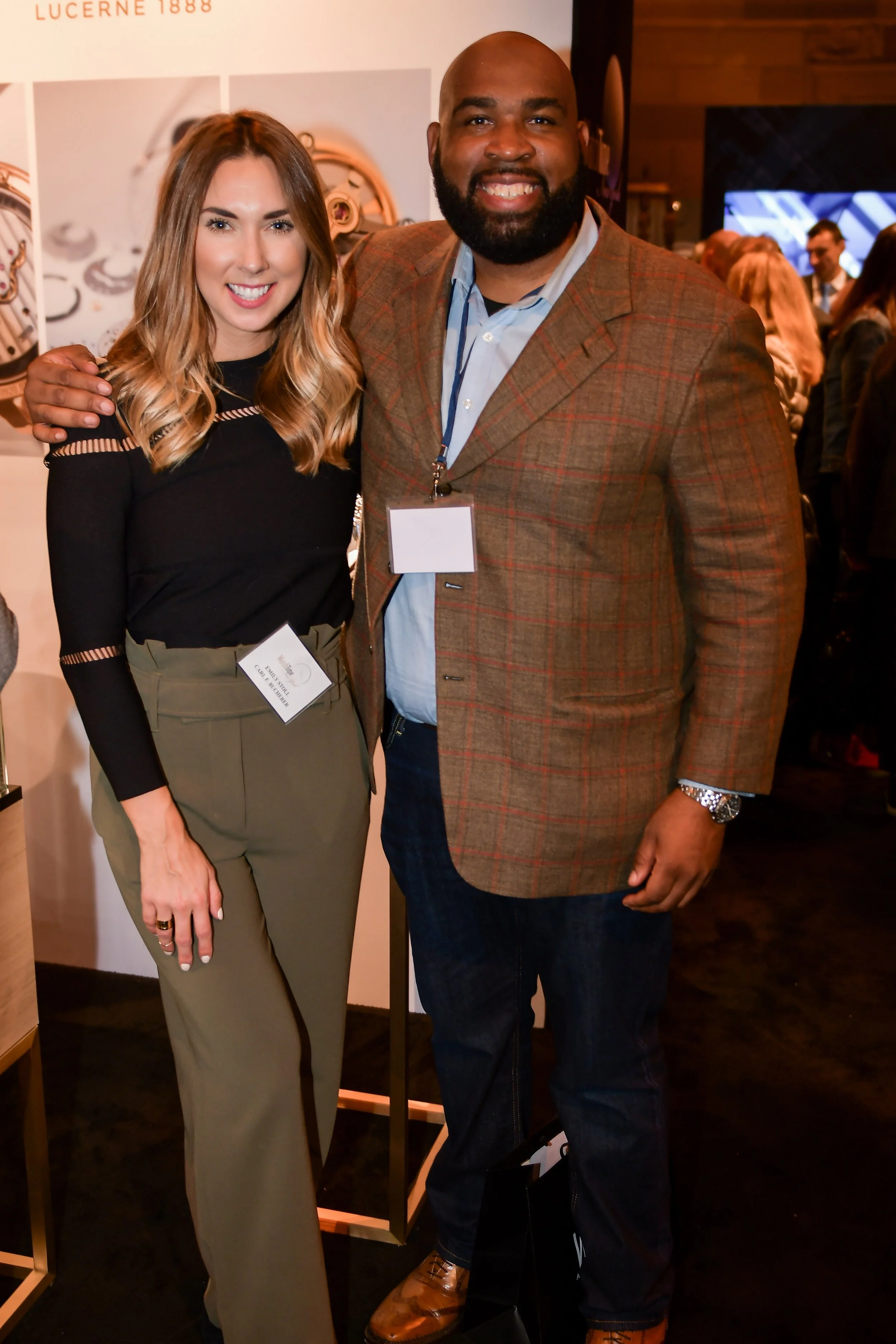 Two people posing together at an event, the woman on the left has long wavy light brown hair, wearing a black top and beige high-waisted pants, and the man on the right has a beard, wearing a brown plaid blazer over a light blue shirt, and holding a 