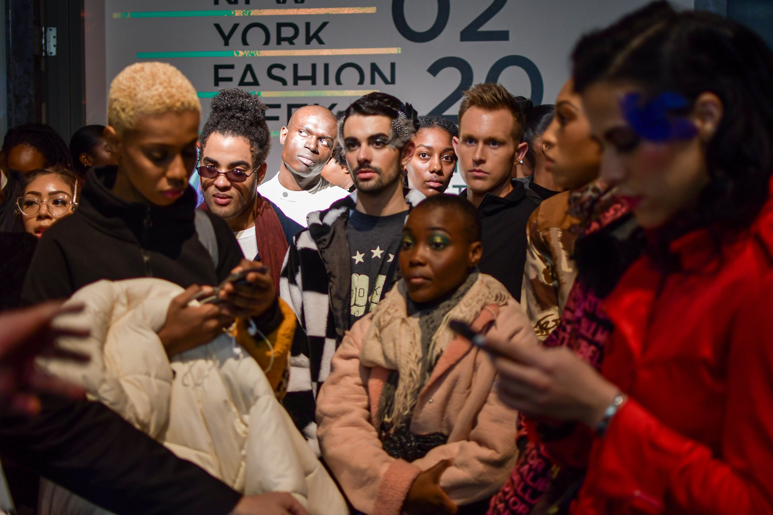 A diverse group of people standing in line, looking at their phones or talking, in front of a sign that says 'NYC YORK 02 FASHION 20'.